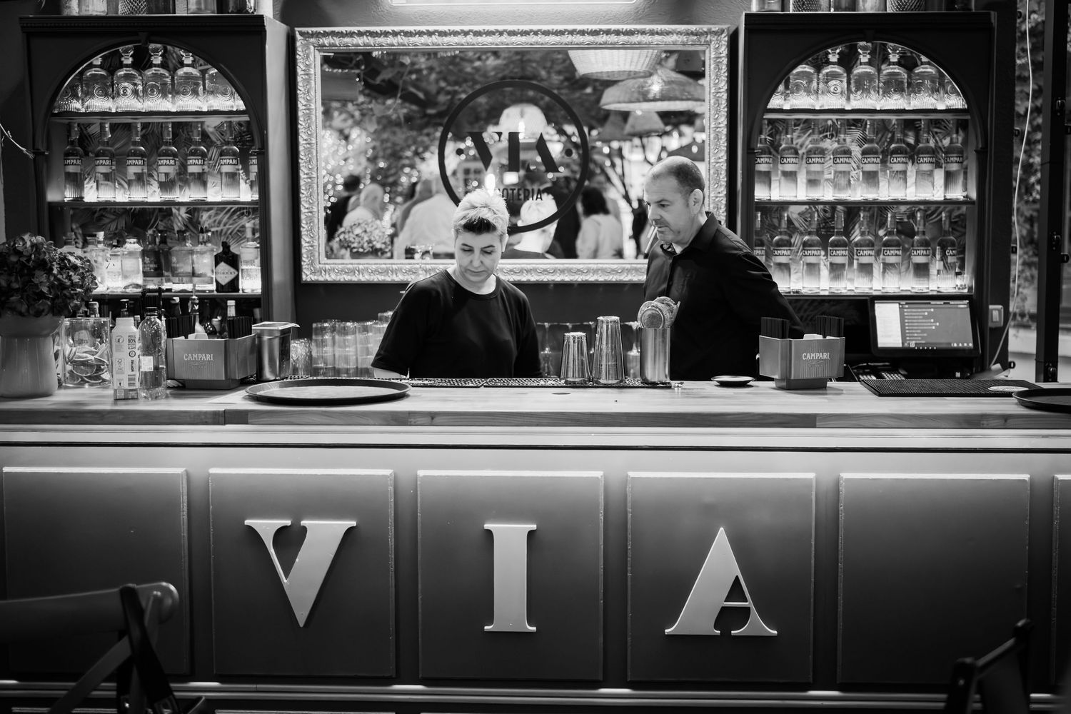 Black and white photo of two men standing behind a bar counter with VIA signage.