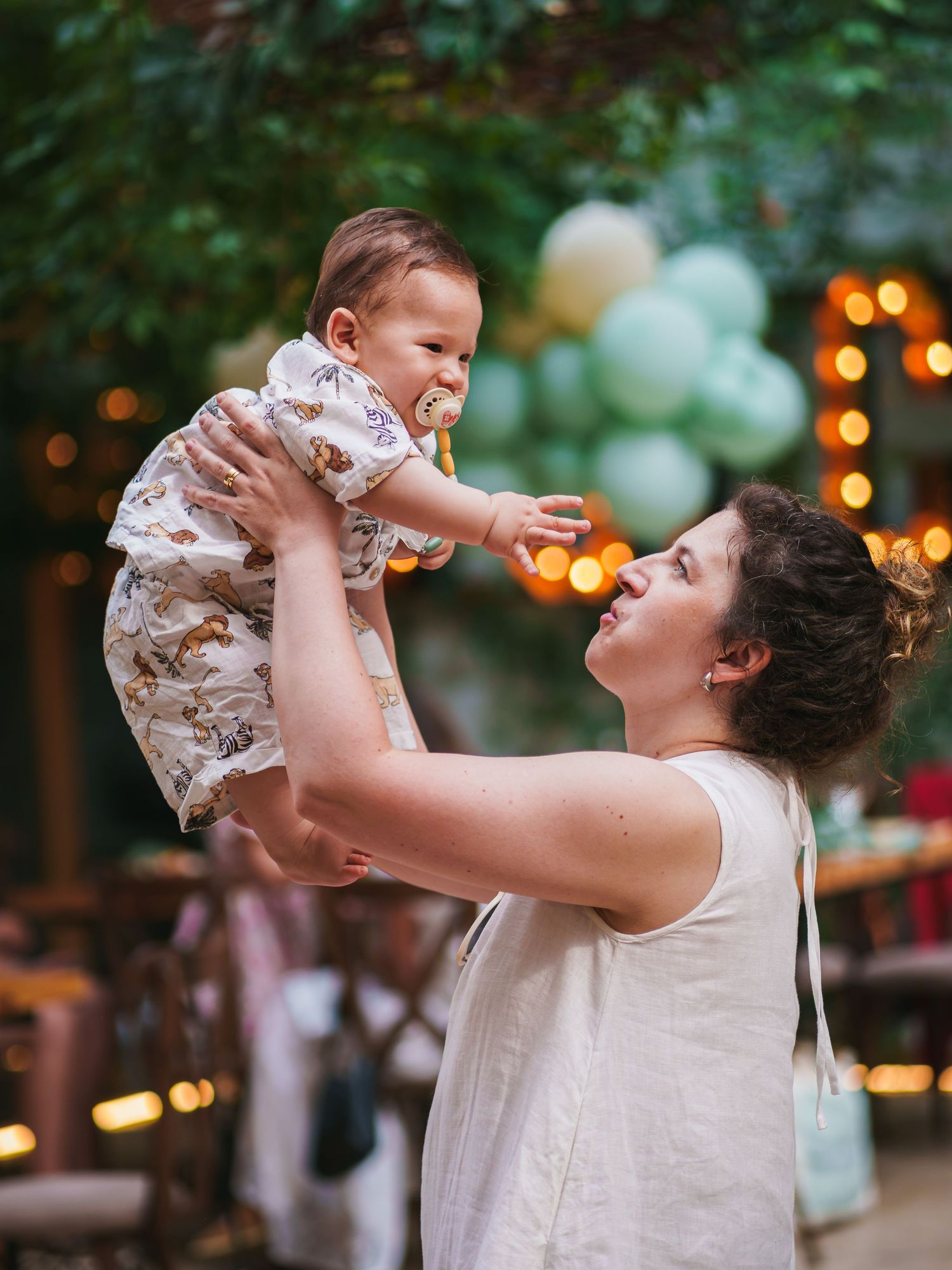 A joyful mother lifts her laughing baby at an outdoor party with green balloons and string lights.