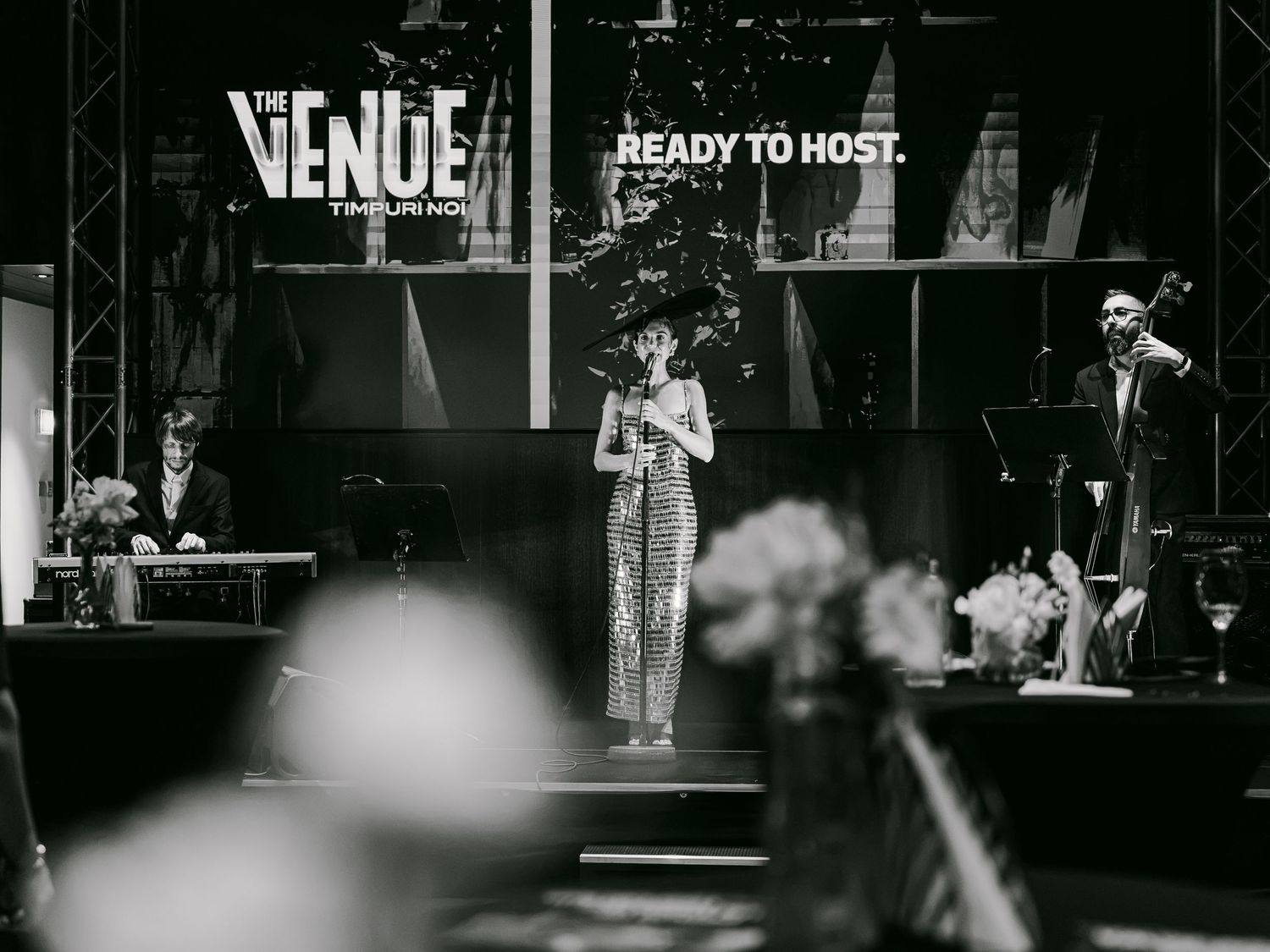Black and white photo of a woman performing on stage at The Venue, with band members and elegant decor visible.