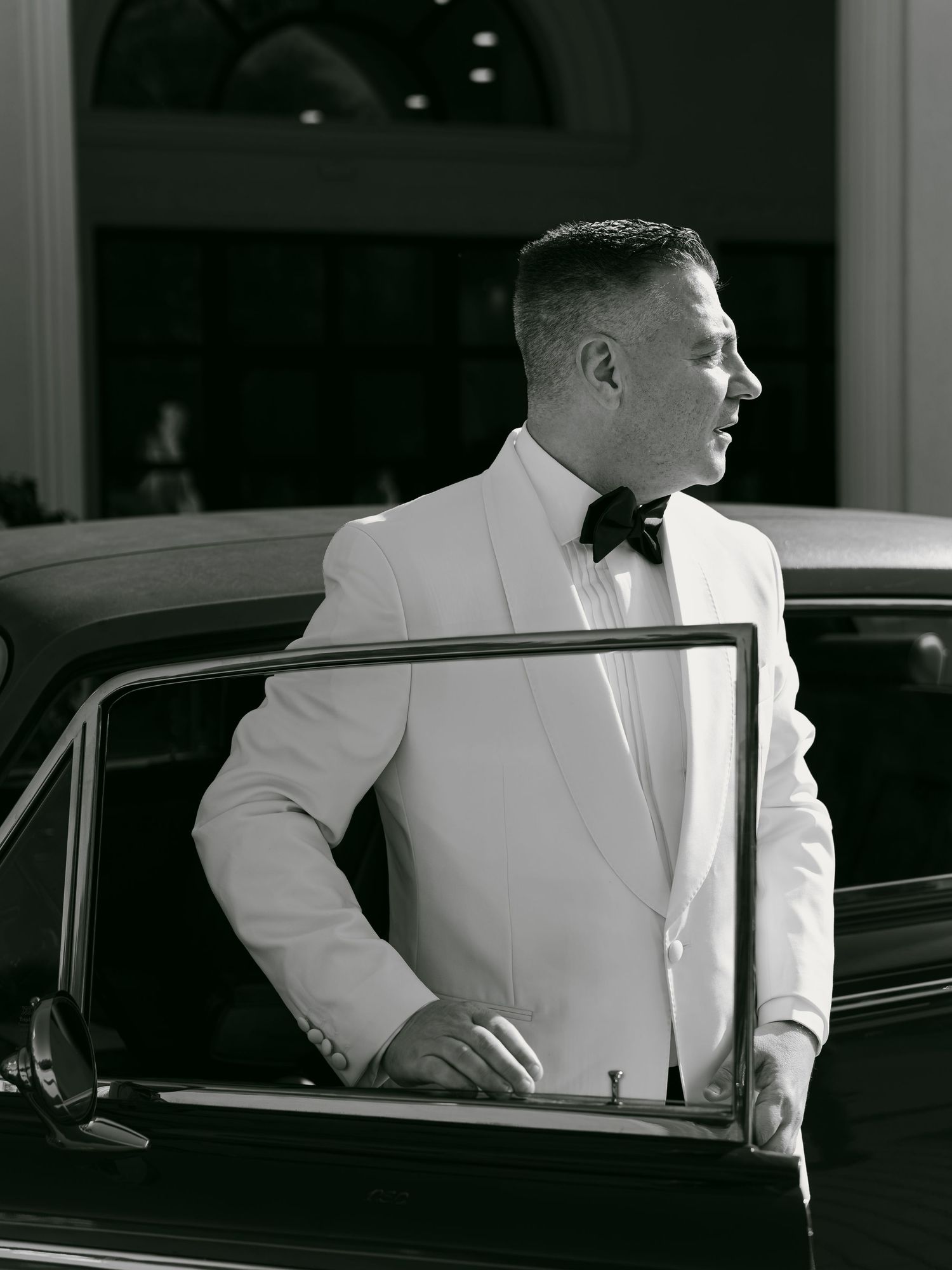 Elegant groom in white tuxedo jacket and black bow tie stepping out of vintage car in black and white photo.