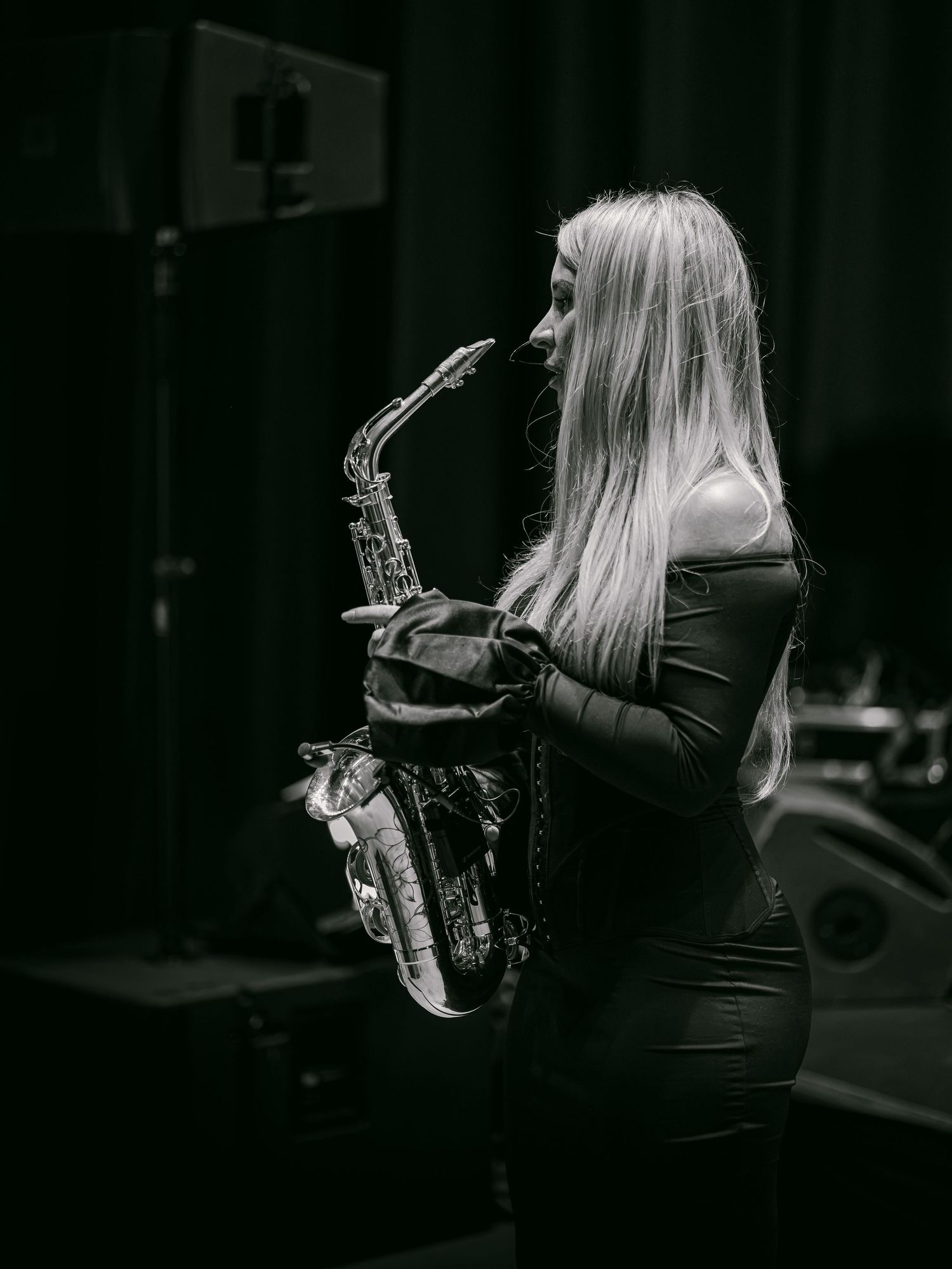 Black and white photo of a blonde woman in a black dress playing a saxophone on a dimly lit stage.