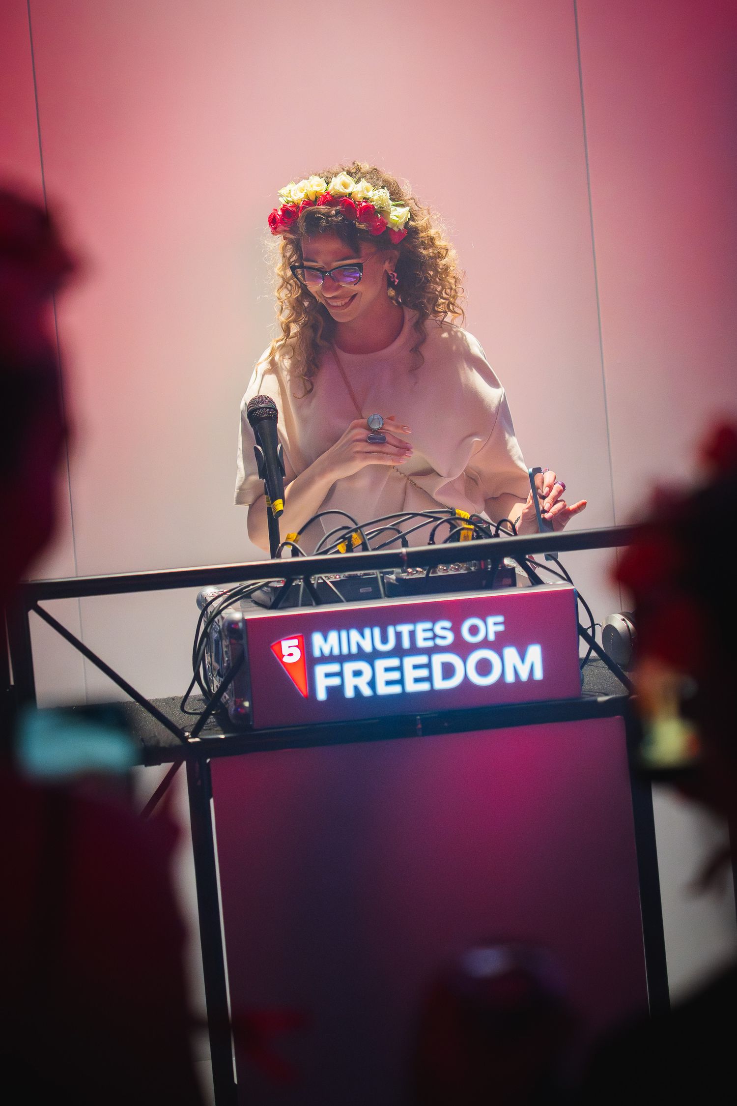 Woman with flower crown DJing at a '5 Minutes of Freedom' event podium under pink stage lighting.