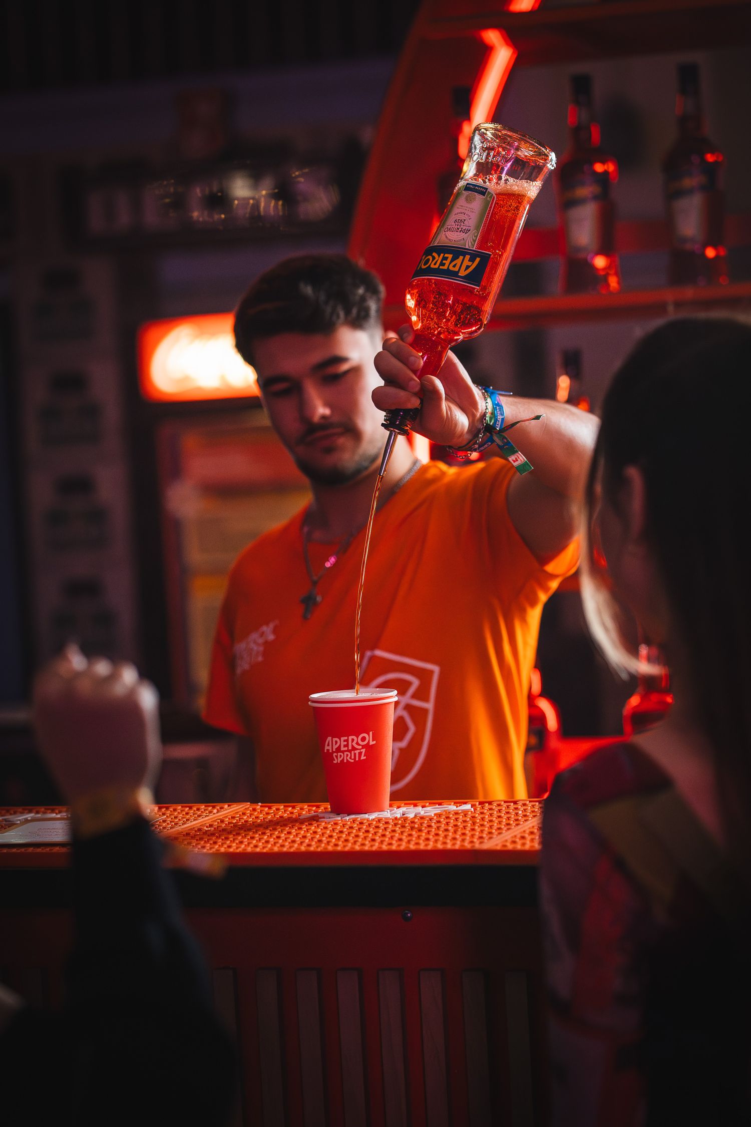Barman in tricou Aperol pregatind un cocktail intr-un bar animat, fotografie comerciala