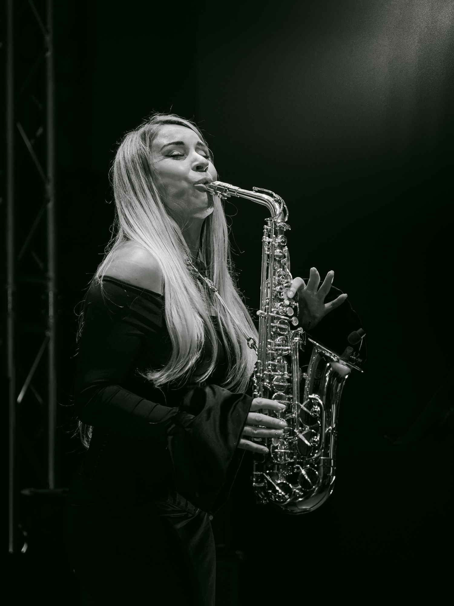 Black and white photo of a blonde woman playing saxophone on stage in dramatic lighting.