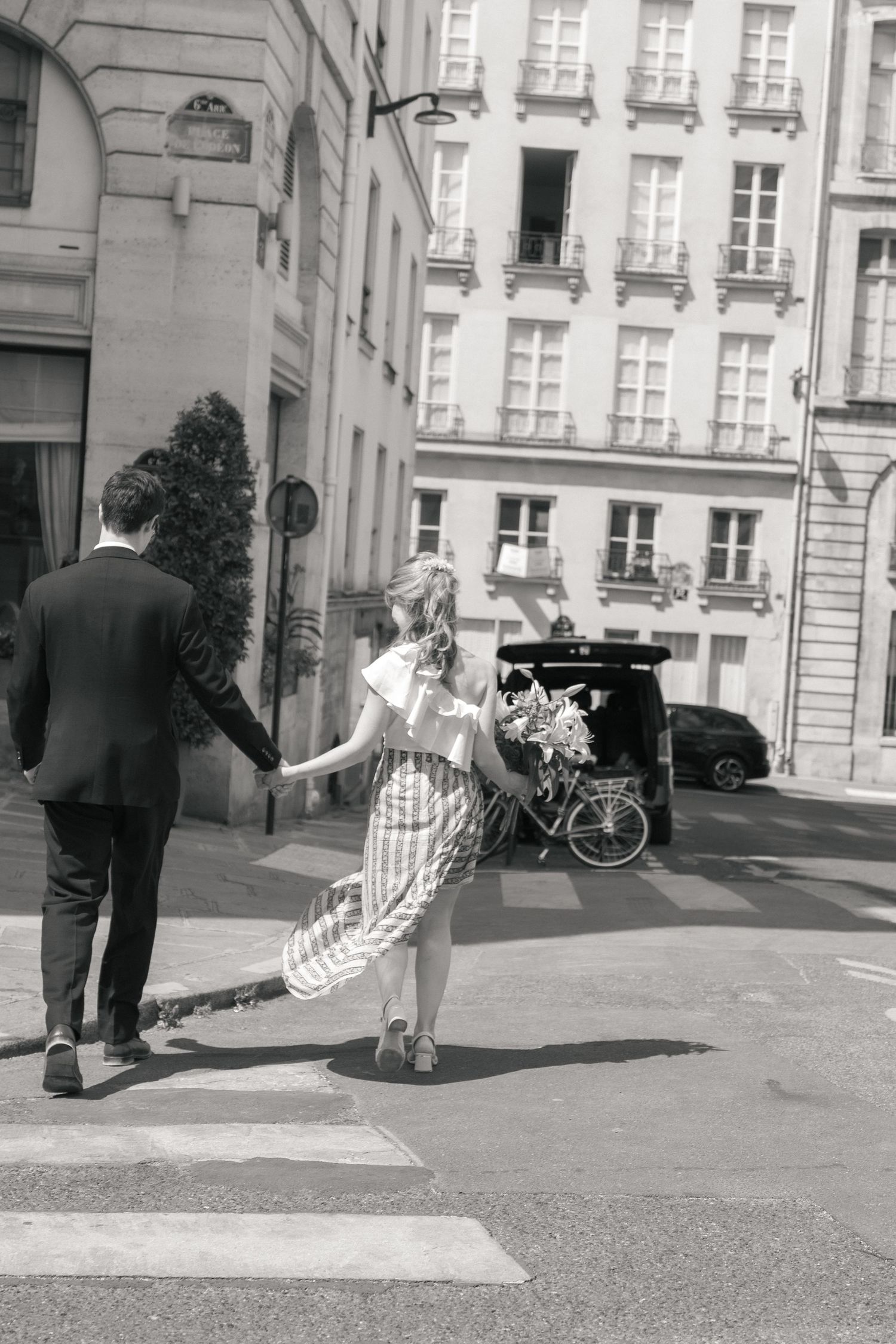 Couple de mariés traversant une rue parisienne main dans la main, photographié en noir et blanc.