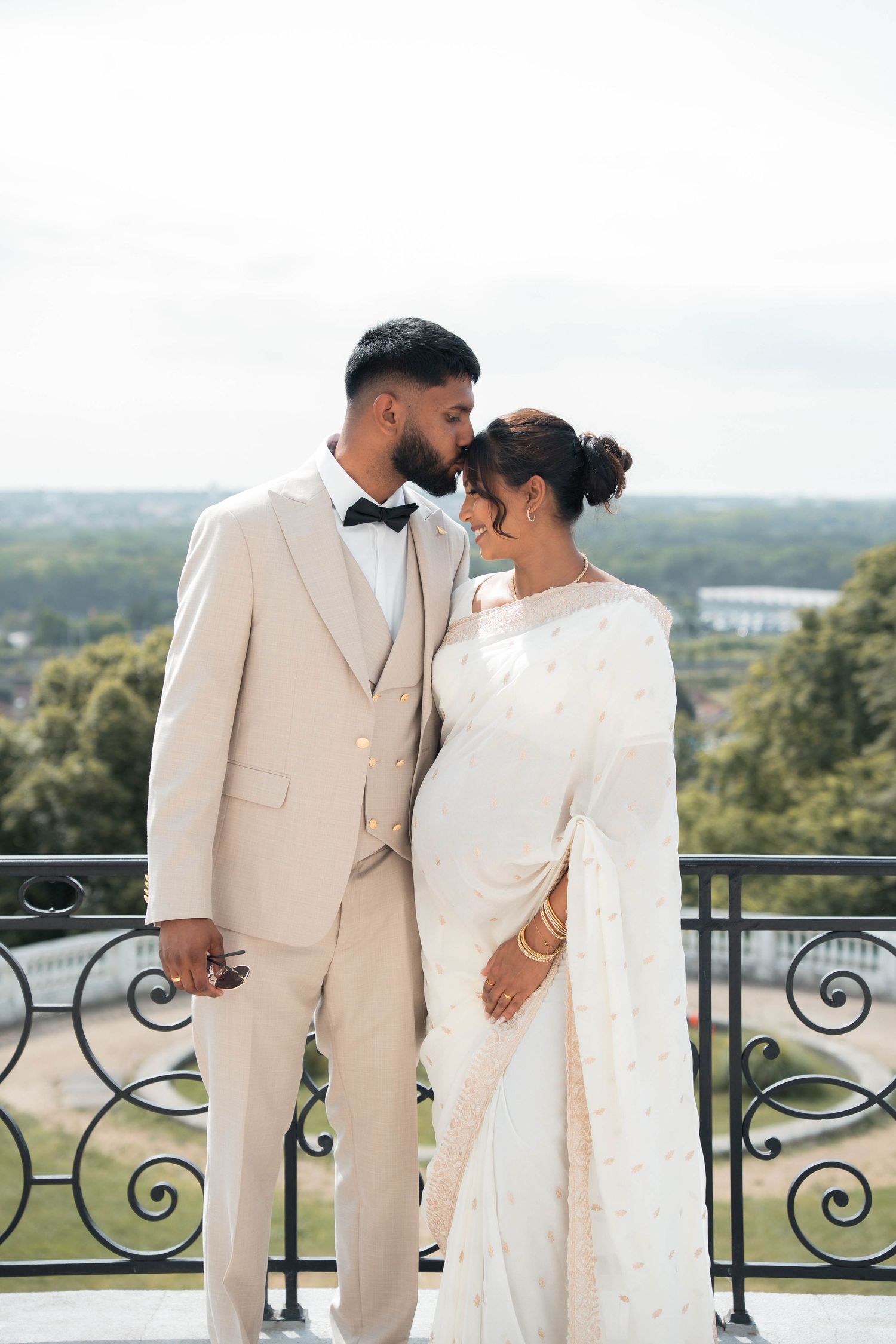 Portrait d’un couple de mariés en tenues claires sur une terrasse dominant le paysage.