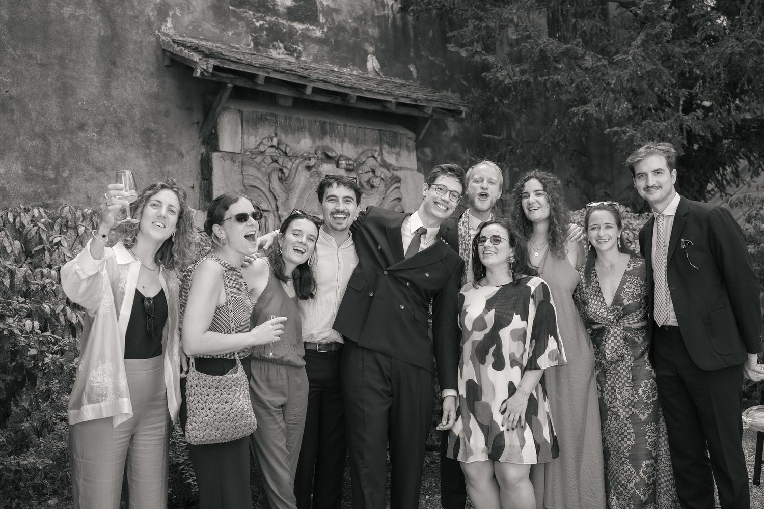 Mariés entourés de leurs amis lors d’une photo de groupe en extérieur, photographiés en noir et blanc.