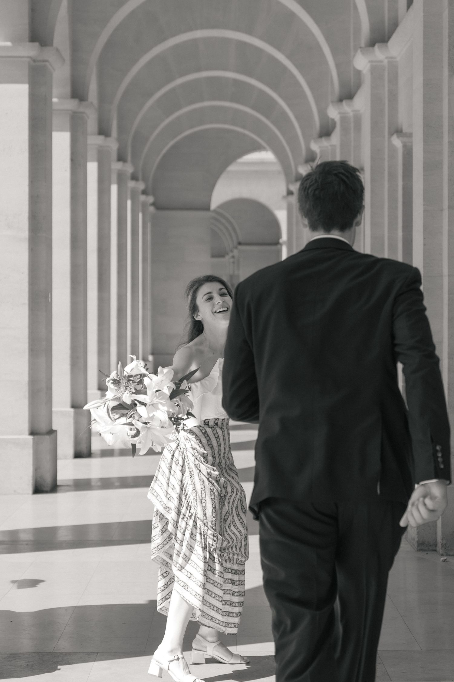 Mariée rejoignant son époux sous une colonnade parisienne après leur cérémonie civile, photographiée en noir et blanc.