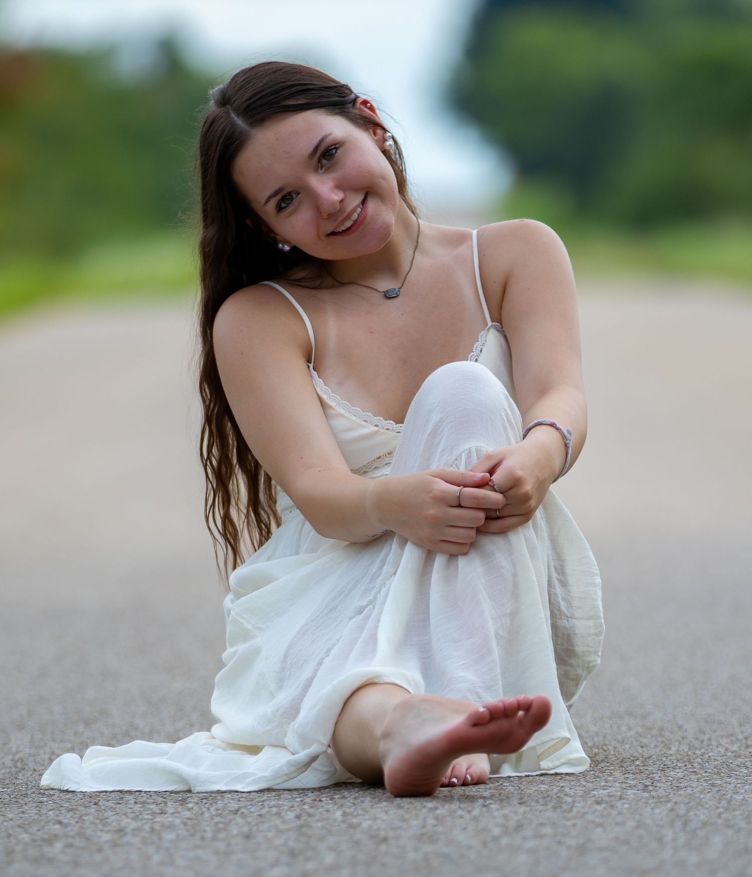 Smiling brunette woman in white dress sitting barefoot on a road with blurred green background.