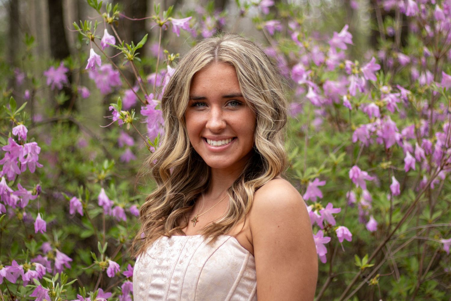 Smiling blonde woman in white lace dress stands among blooming purple azalea bushes outdoors.