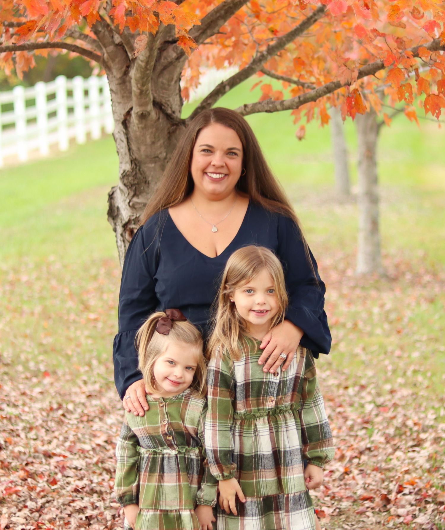 Smiling mother poses with two young daughters in matching plaid dresses under autumn tree.