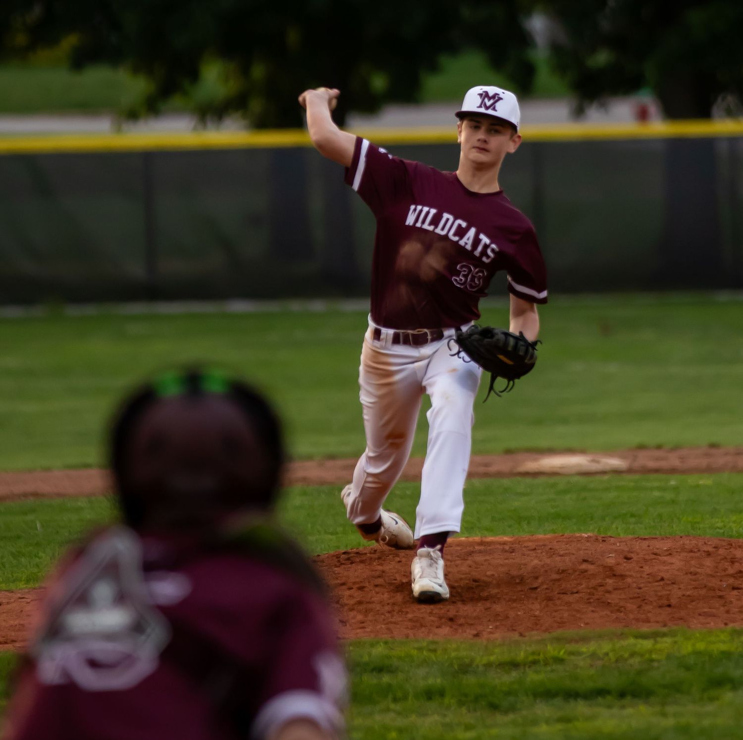 Wildcats baseball pitcher in maroon uniform throwing ball toward home plate.