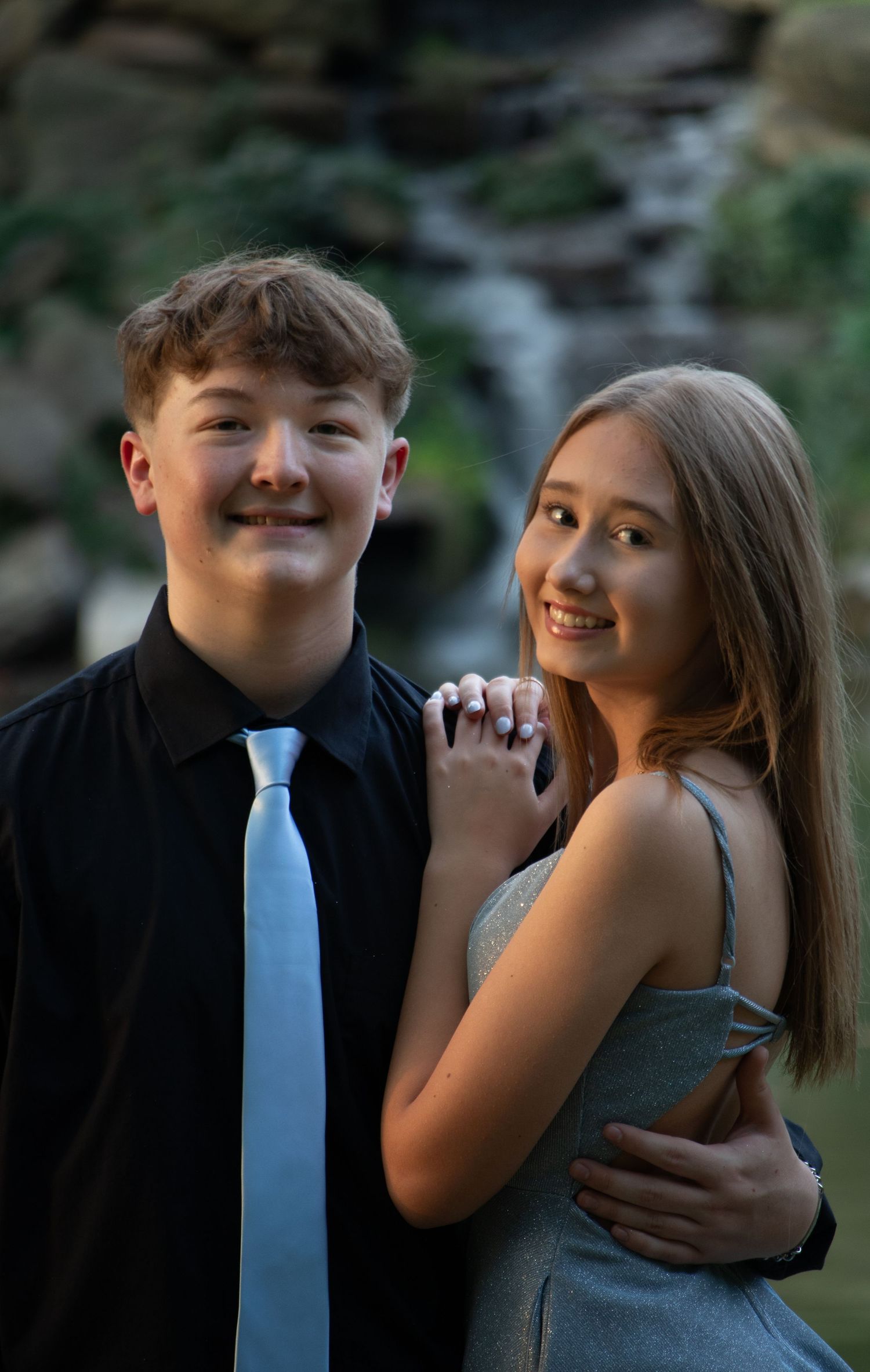 Smiling teenage couple posing together in front of scenic waterfall backdrop.