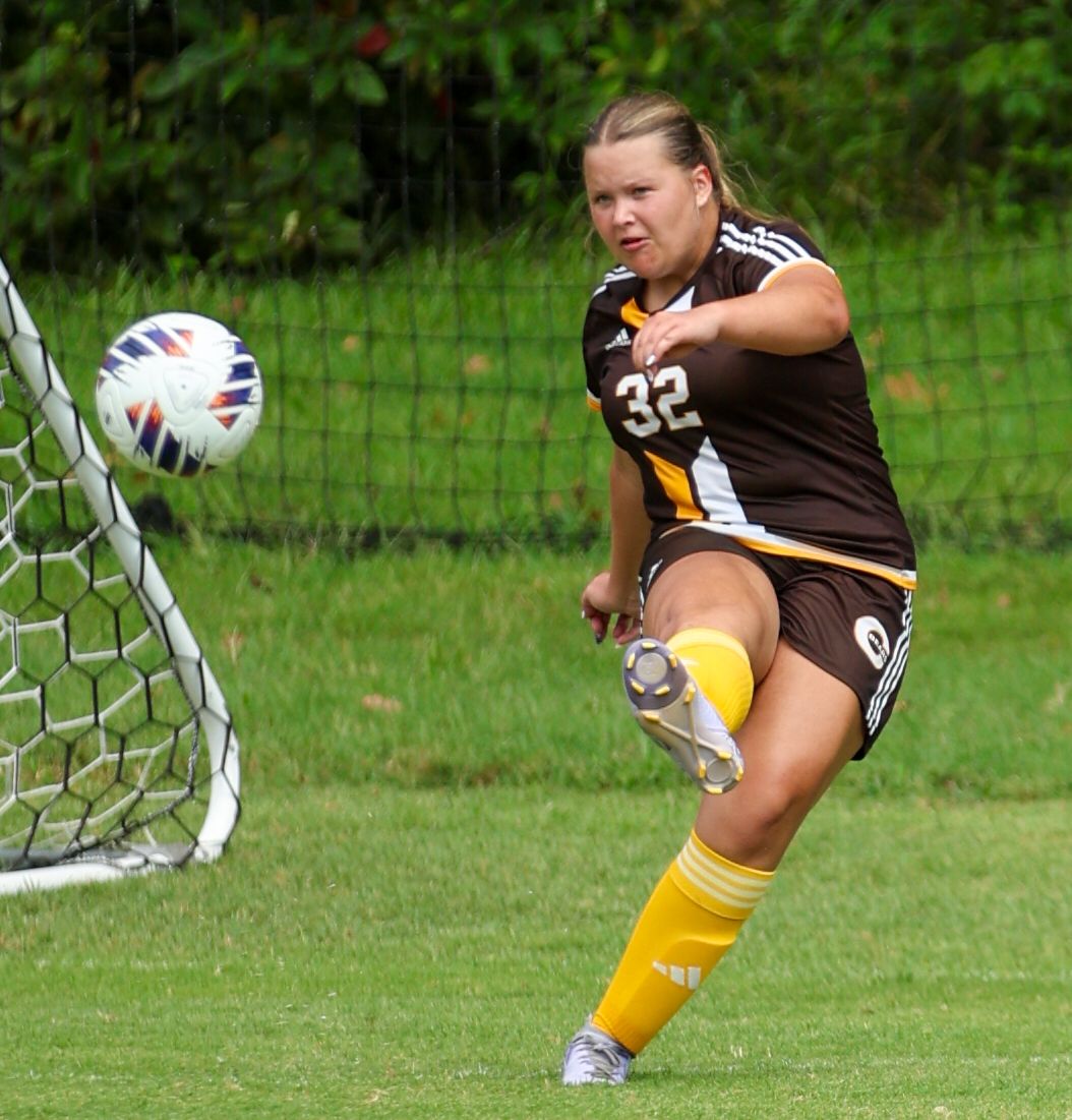 Female soccer player #32 in brown uniform kicking ball near goal on green field.
