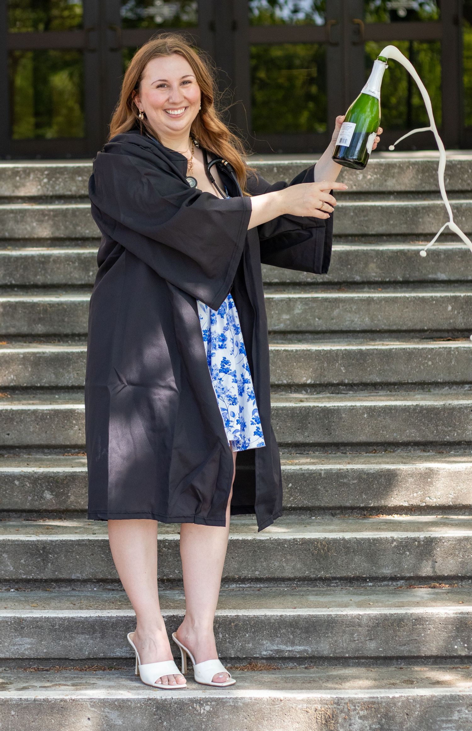 Woman in graduation gown celebrating with champagne bottle on outdoor steps.