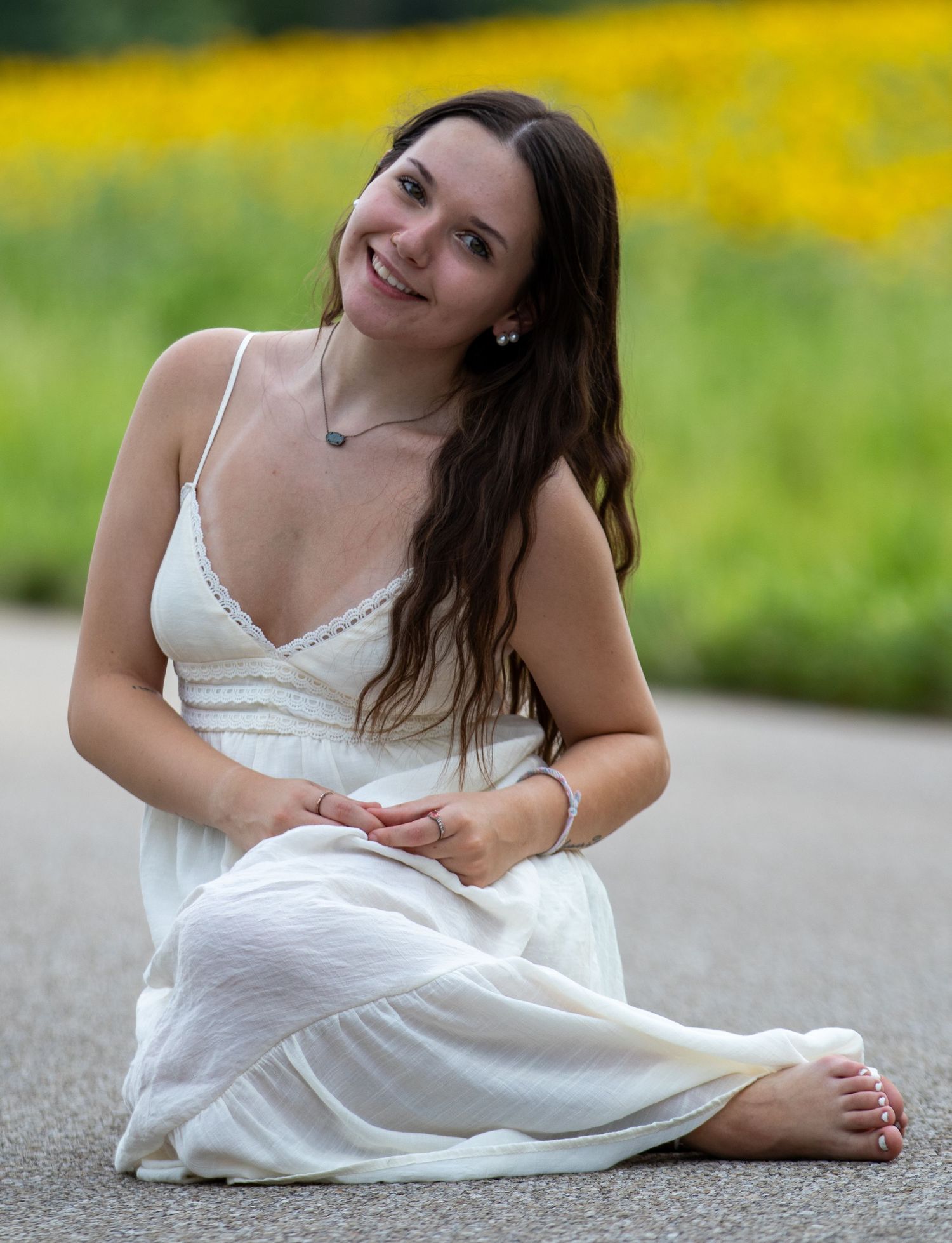 Cheerful young woman in white sundress sitting barefoot on road with yellow flowers in background.