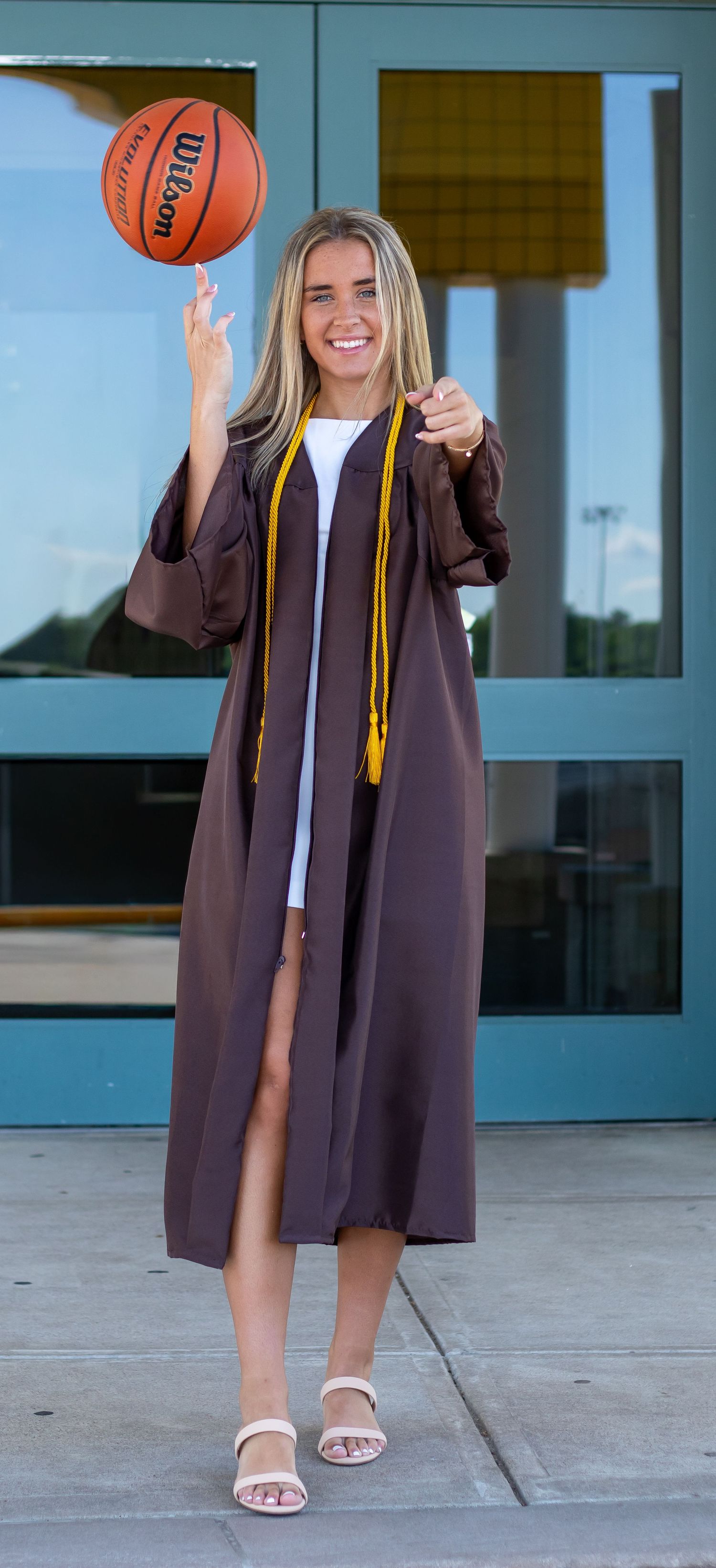 Blonde graduate in brown gown spinning a Wilson basketball on her finger outdoors.