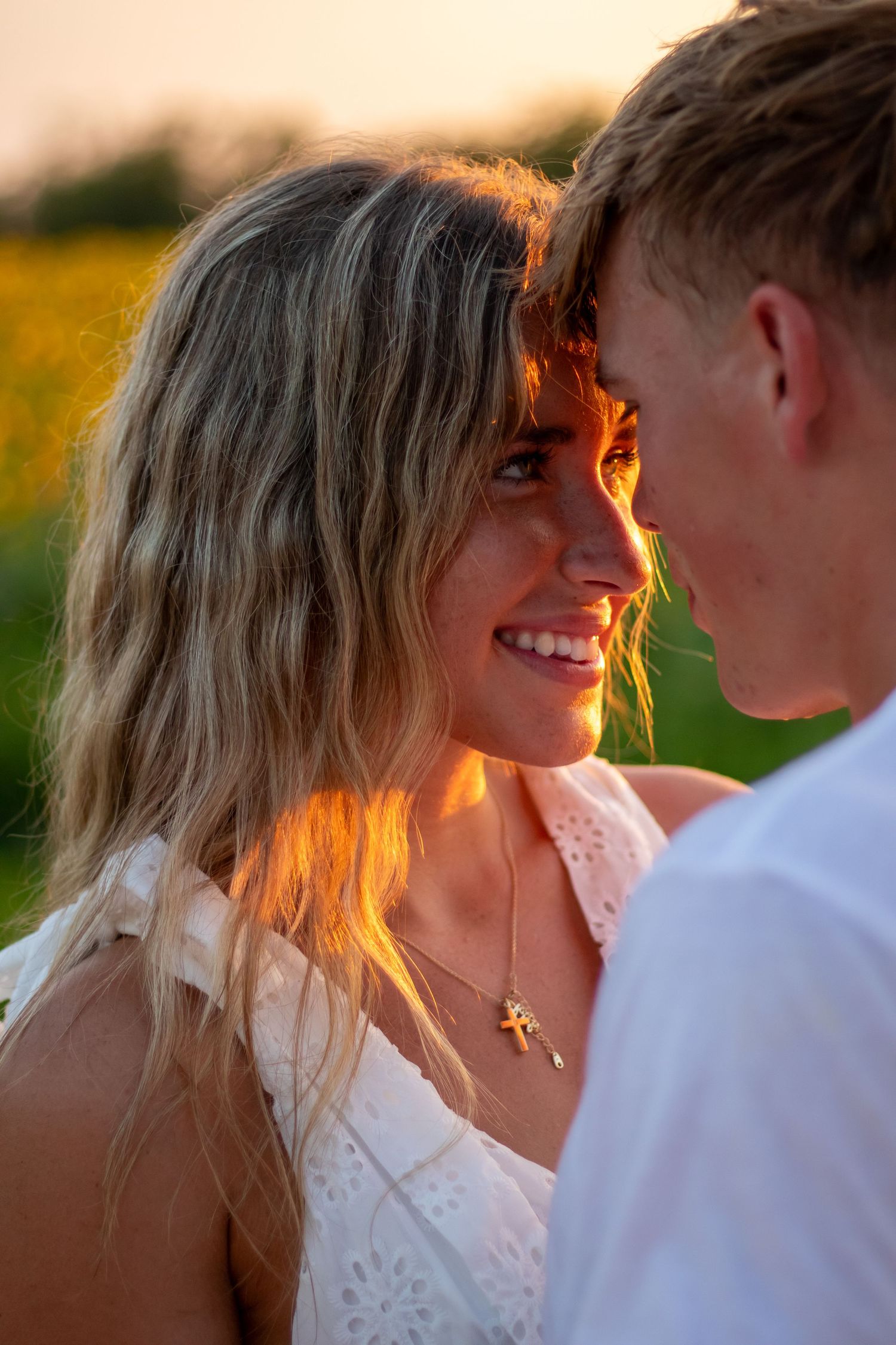Smiling blonde woman in white dress with golden sunset light illuminating her face in a sunflower field.