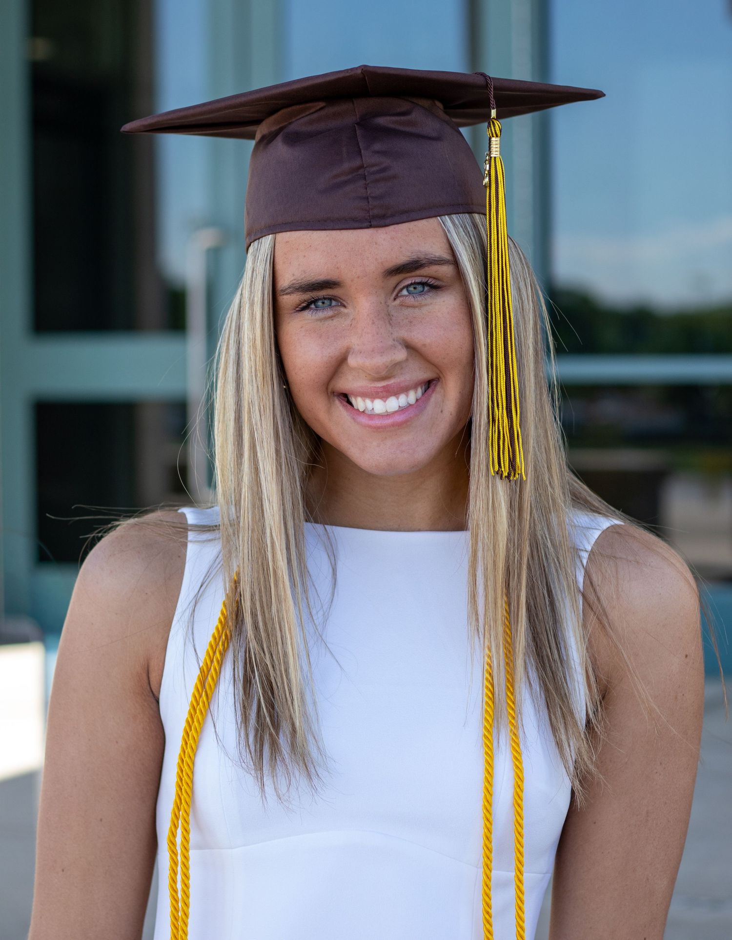Close-up of smiling blonde graduate wearing brown mortarboard cap with gold tassel outdoors.