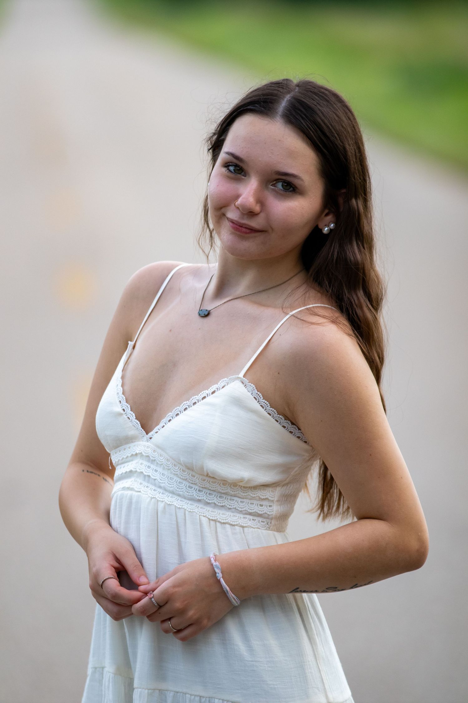 Young woman in white sundress poses on a road, smiling softly with long dark hair flowing over her shoulder.