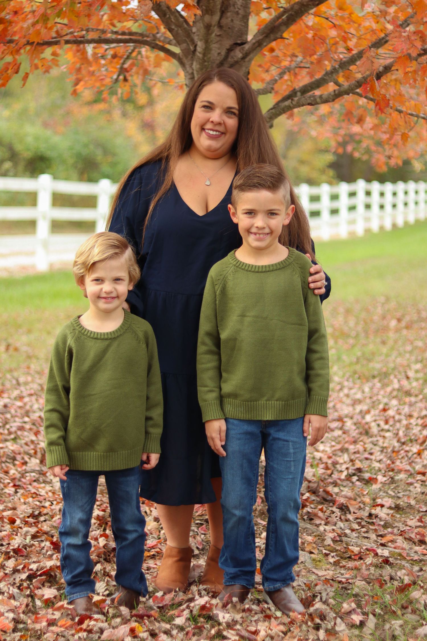 Happy mother smiles with two young boys in matching green sweaters under vibrant fall foliage.