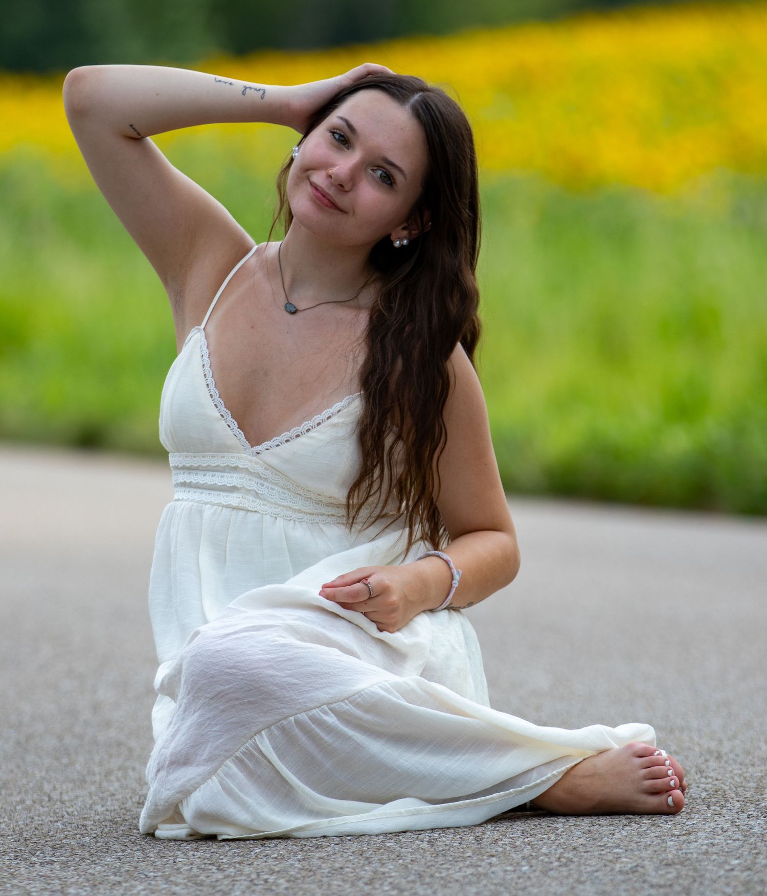 Young woman in white sundress sitting barefoot on pavement with yellow wildflowers in background.