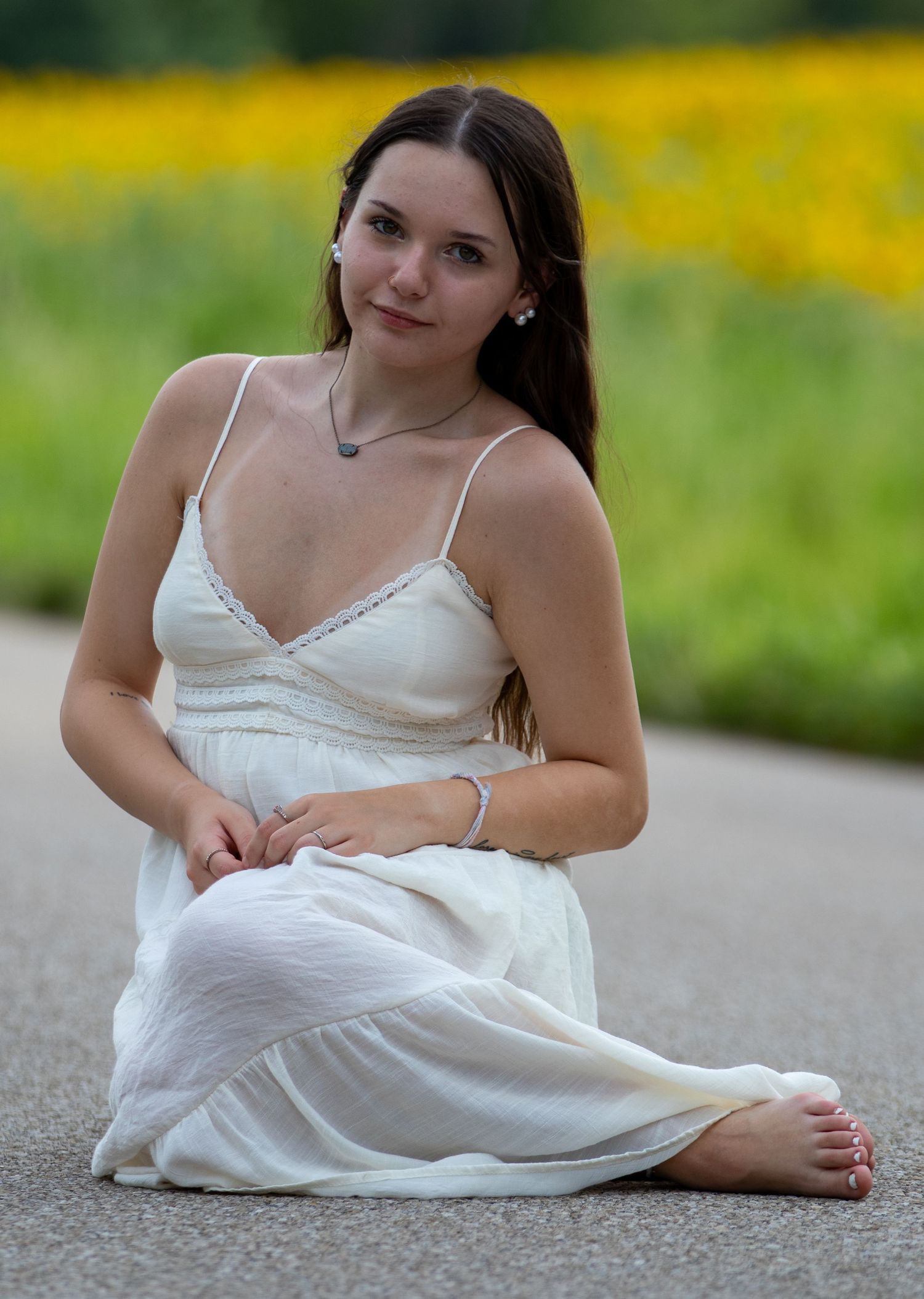 Young brunette woman in white spaghetti strap dress sitting barefoot on a road near a flower field.