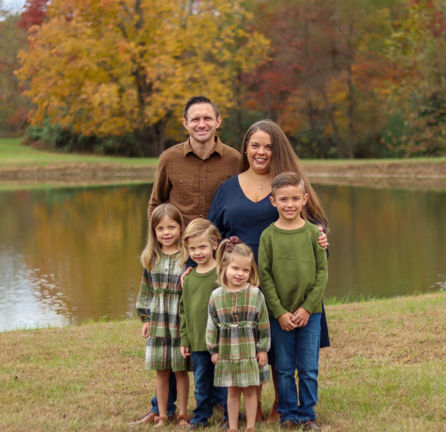 Family of six posing near autumn pond with colorful fall foliage background.
