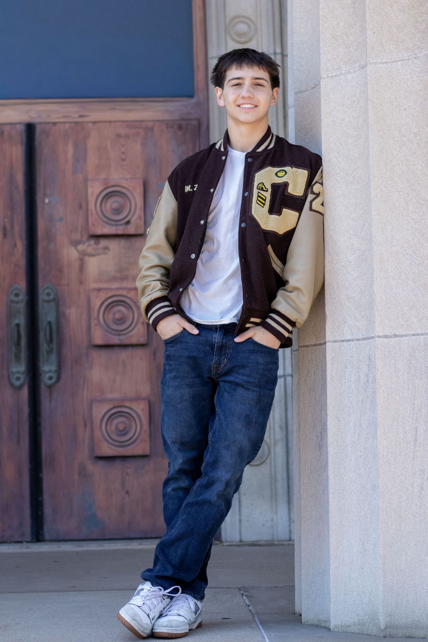 Teenage boy in brown and tan varsity jacket leaning against stone wall by wooden doors.