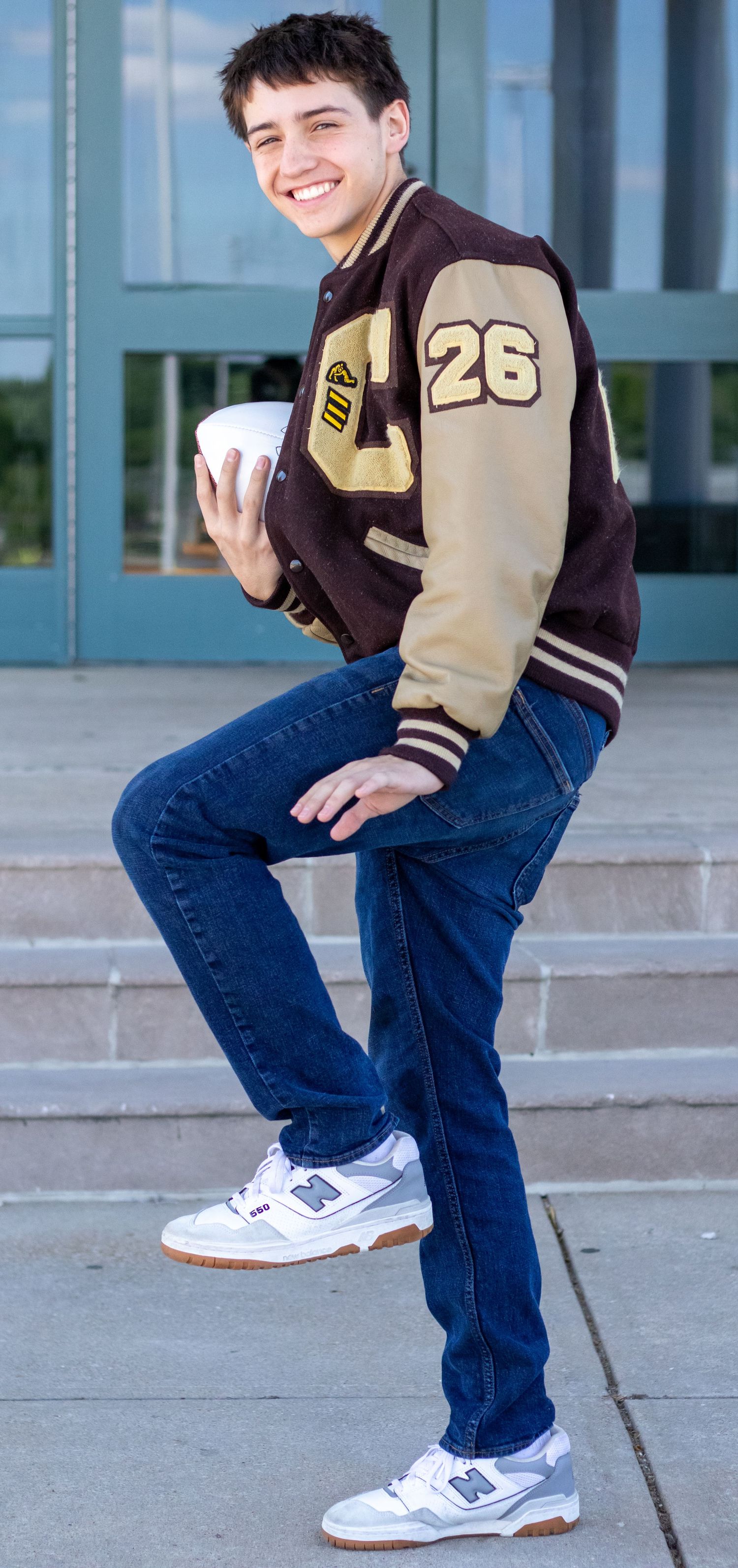 Teen boy in varsity jacket number 26 posing on school steps holding a white cup.