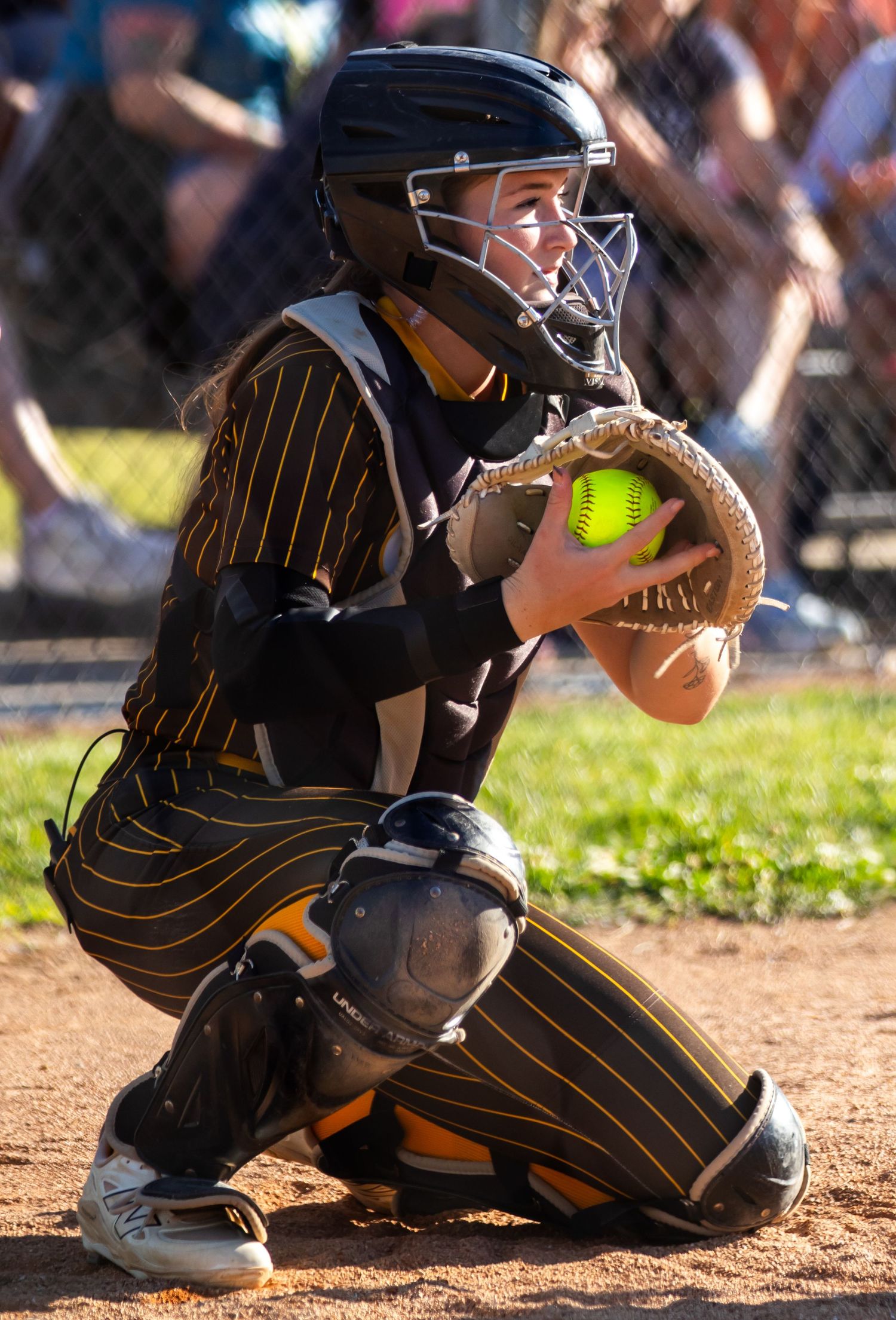 Central softball catcher in full gear crouches and catches a yellow softball during a game.