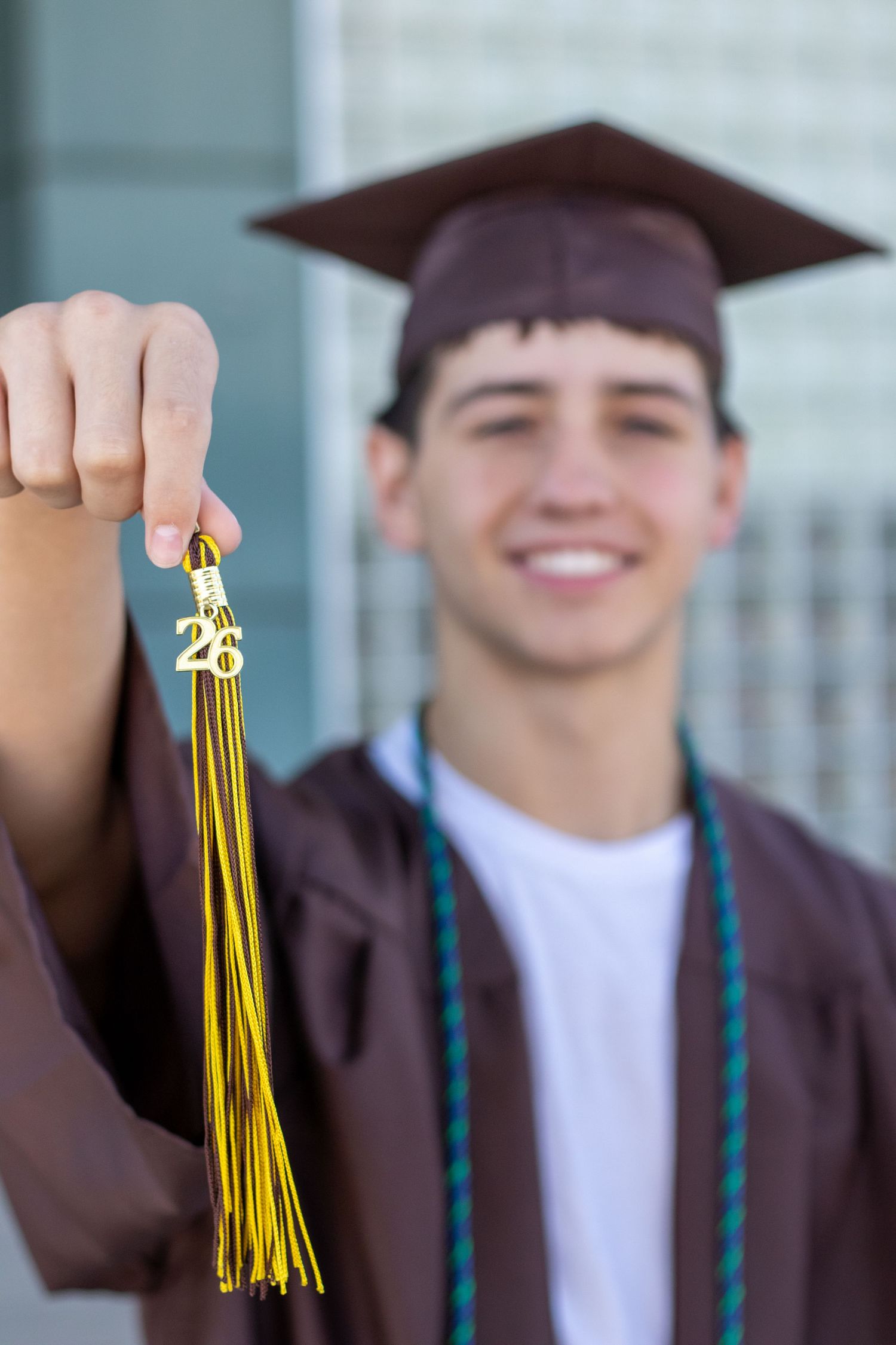 High school graduate in brown cap and gown holding gold tassel with 26 charm closeup.