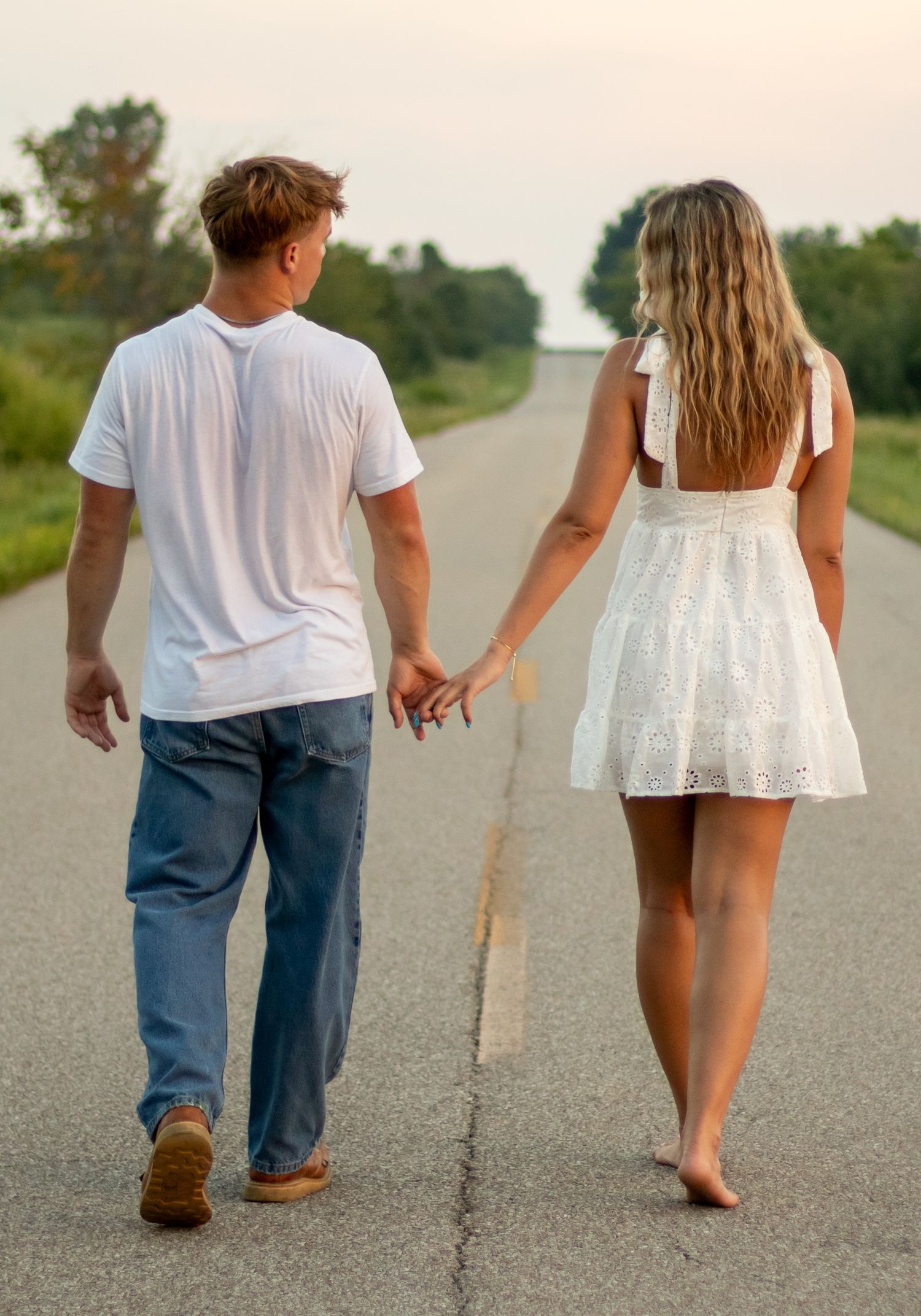 Couple walking hand in hand down a long straight road surrounded by green trees at dusk.