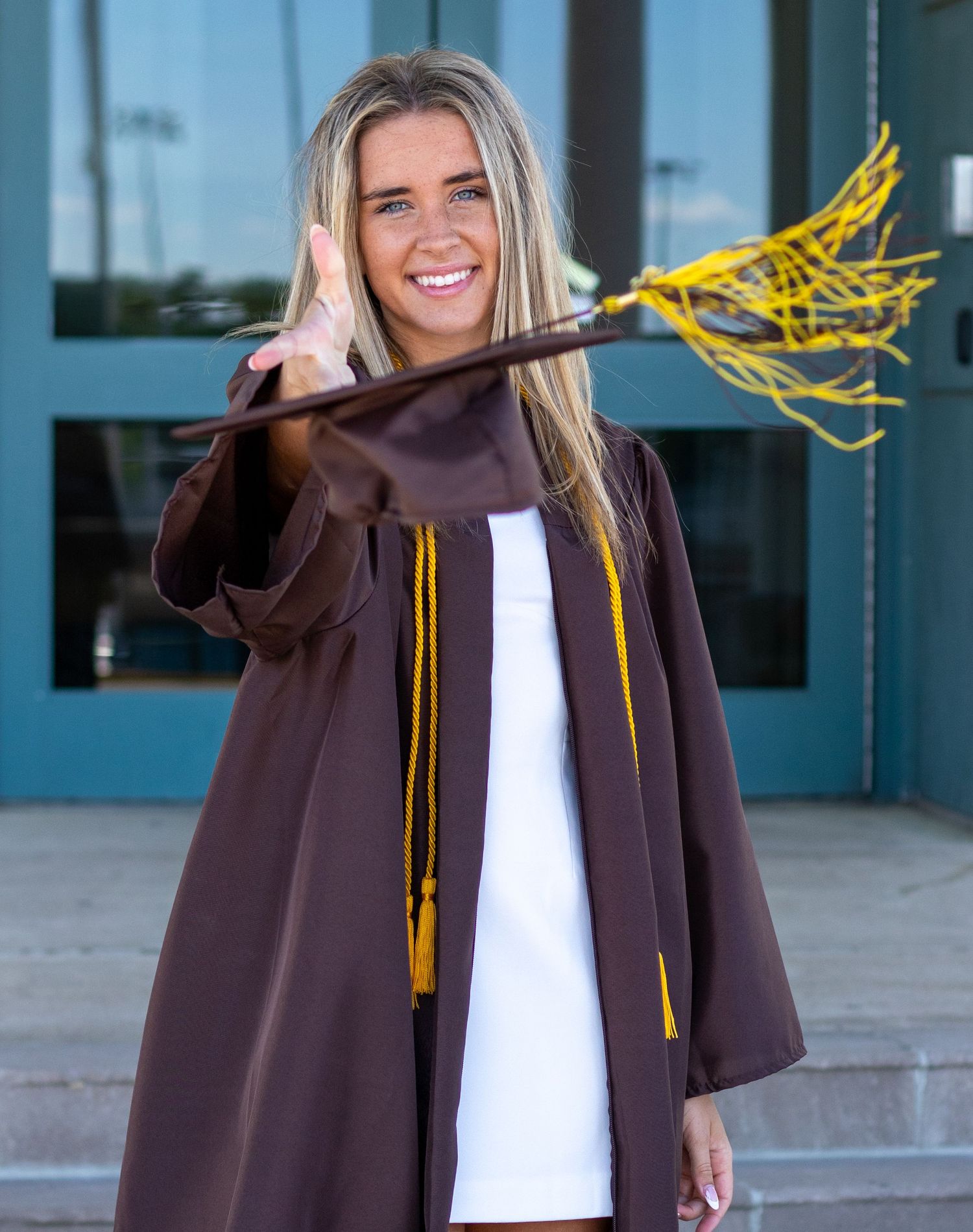 Smiling blonde graduate in brown gown holding a lacrosse stick with gold tassels.