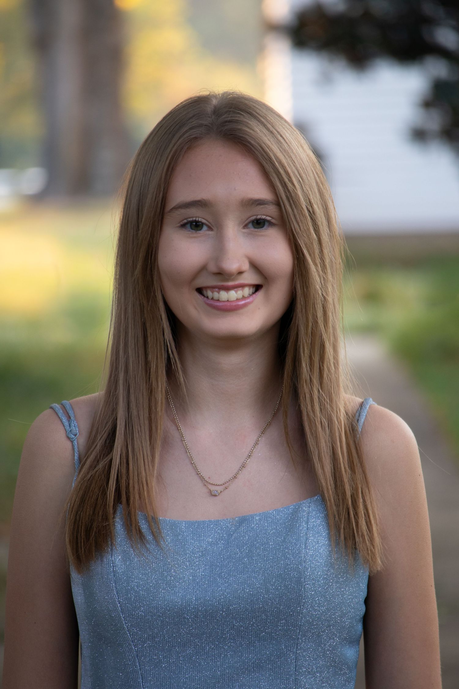 Smiling teenage girl with straight blonde hair wears a blue sparkly dress outdoors in soft light.