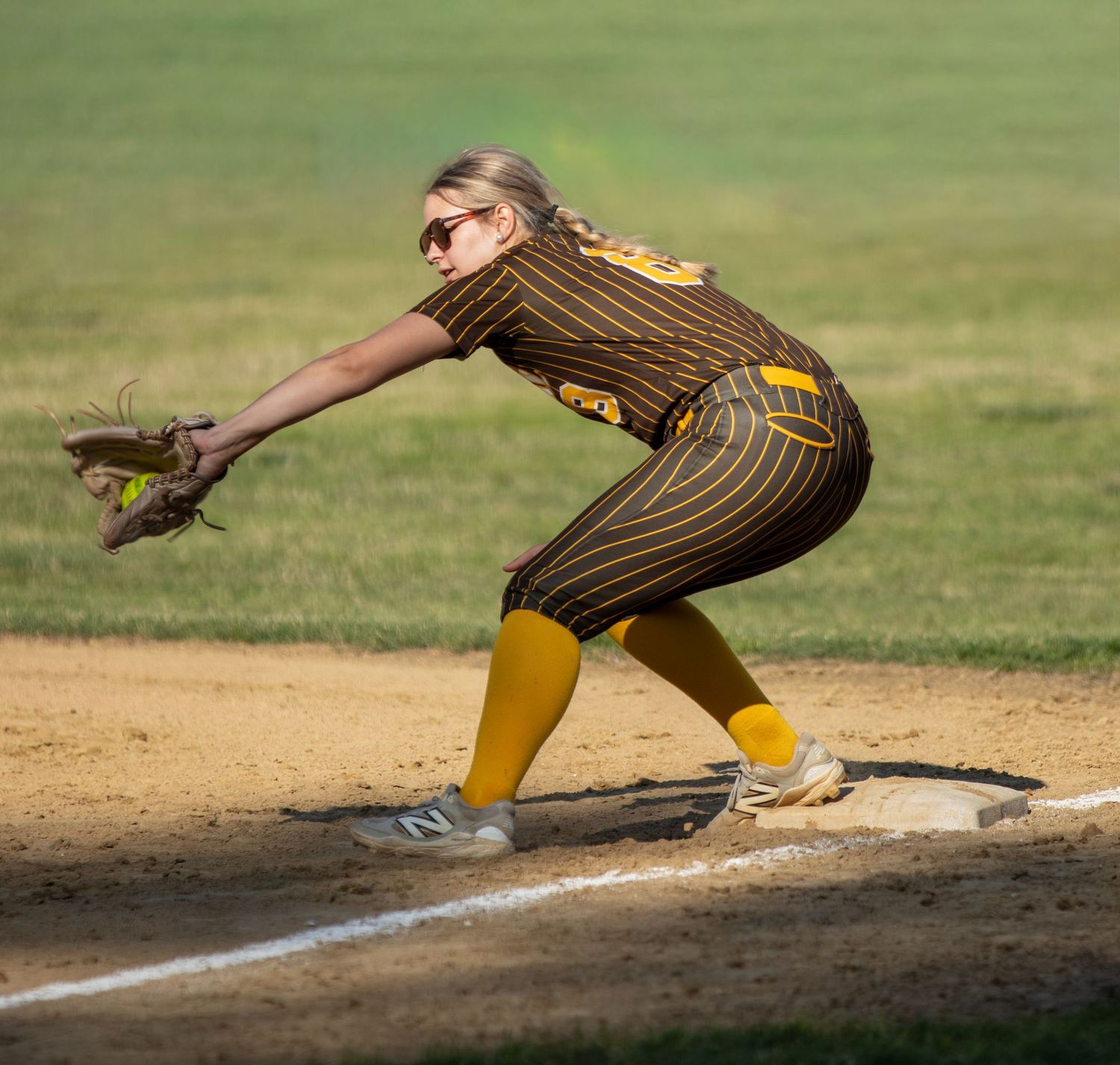 Female softball player in brown pinstripe uniform fielding a ball at first base during a sunny game.