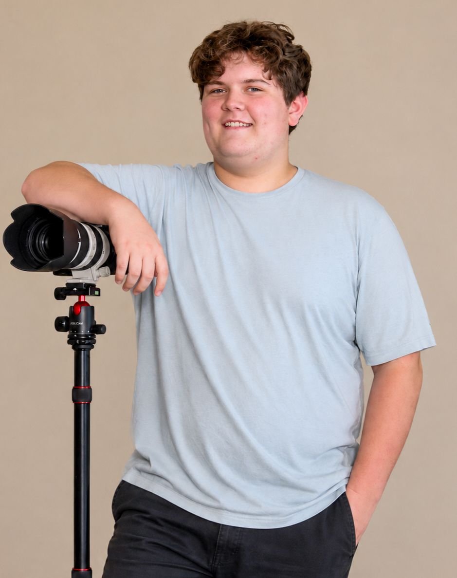 Young male photographer smiling while leaning on professional camera tripod.