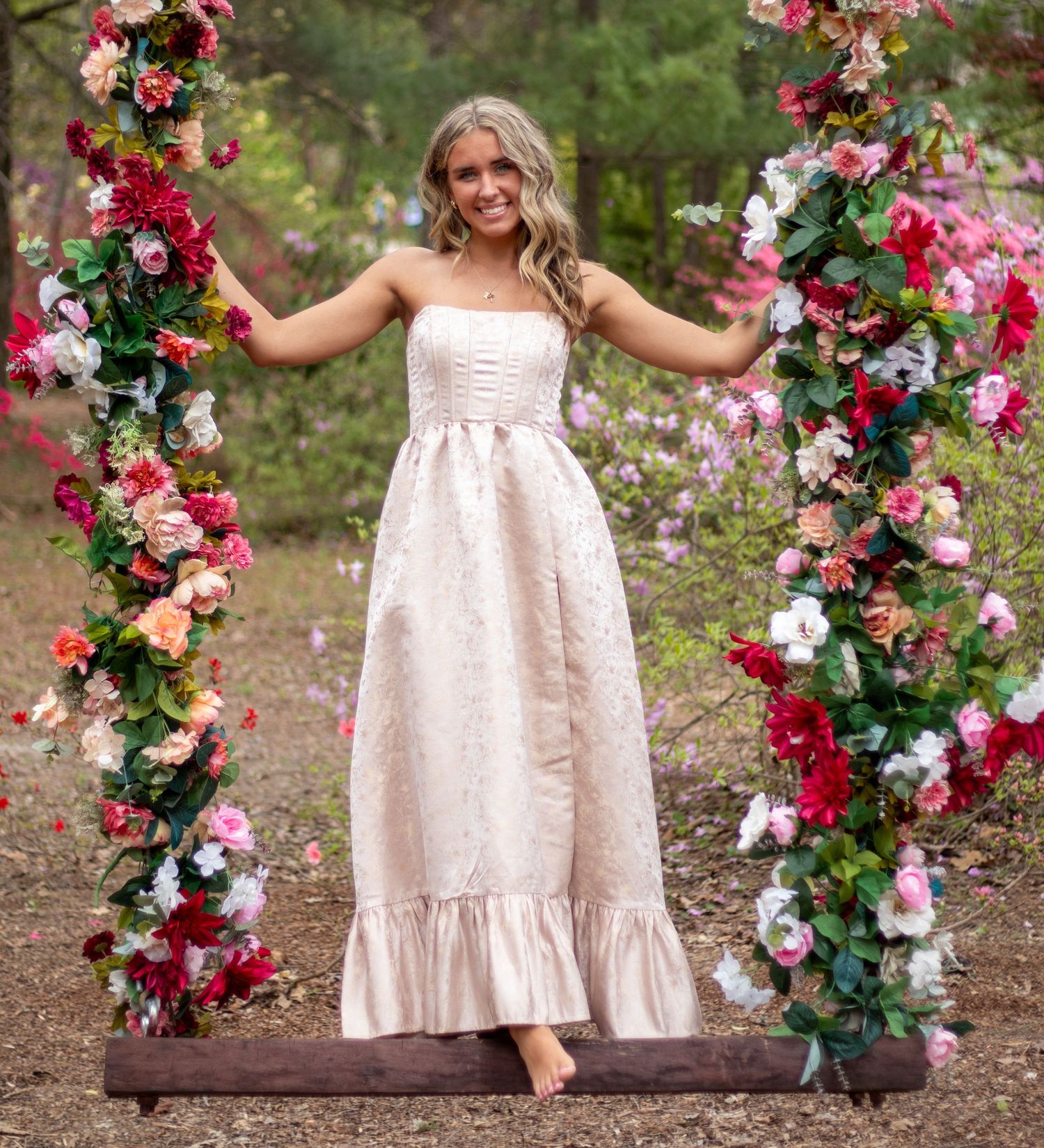 Blonde woman in strapless pink floral gown stands barefoot on a flower-decorated wooden swing outdoors.