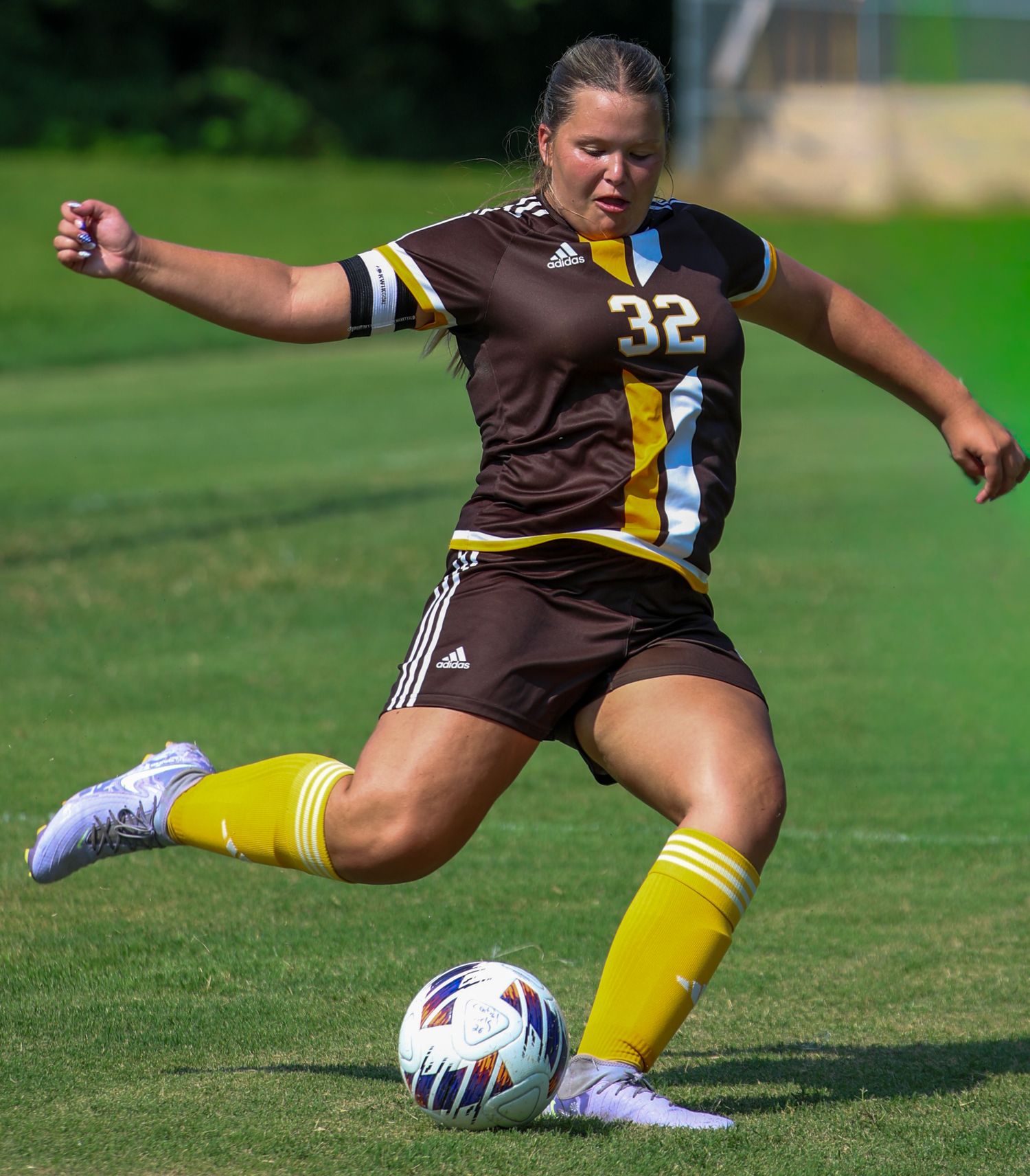 Female soccer player wearing number 32 brown and gold uniform kicking a soccer ball on a green grass field.