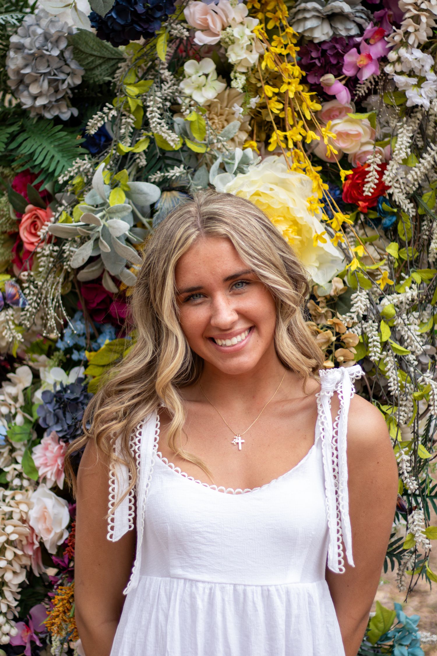 Smiling blonde woman in white sundress poses before a vibrant colorful floral wall backdrop.