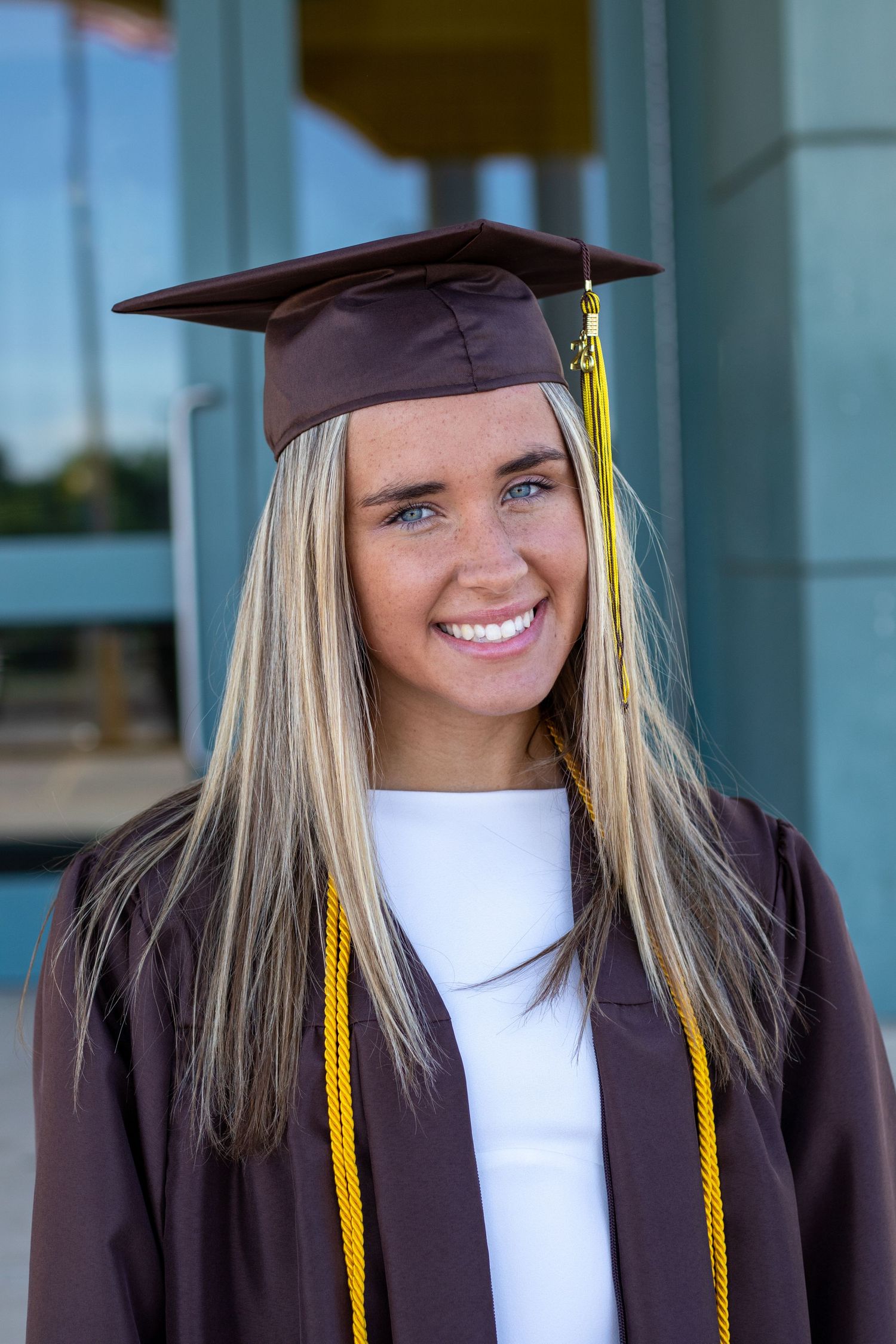 Smiling blonde graduate with blue eyes wearing brown cap and gown with gold honor cord.