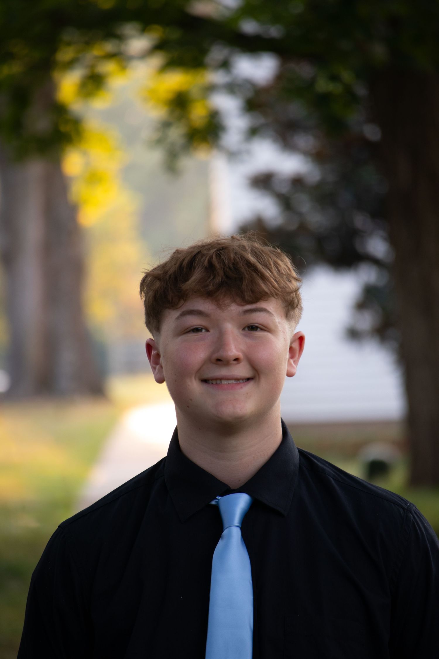 Smiling teenage boy in black shirt and light blue tie poses outdoors with trees in background.