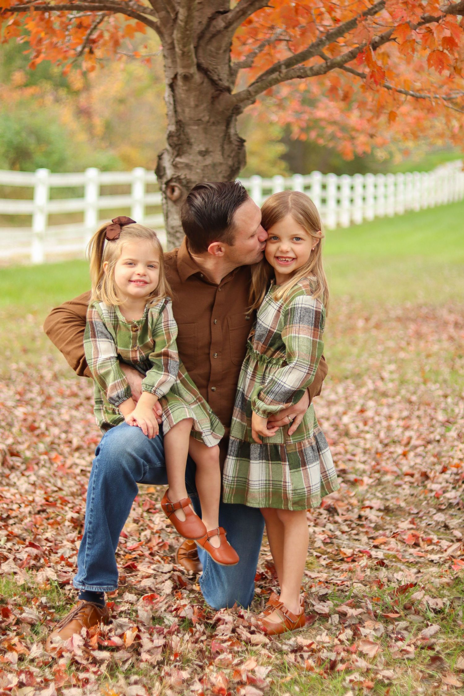 Father kneels kissing daughter while two girls in matching plaid dresses pose under autumn tree.