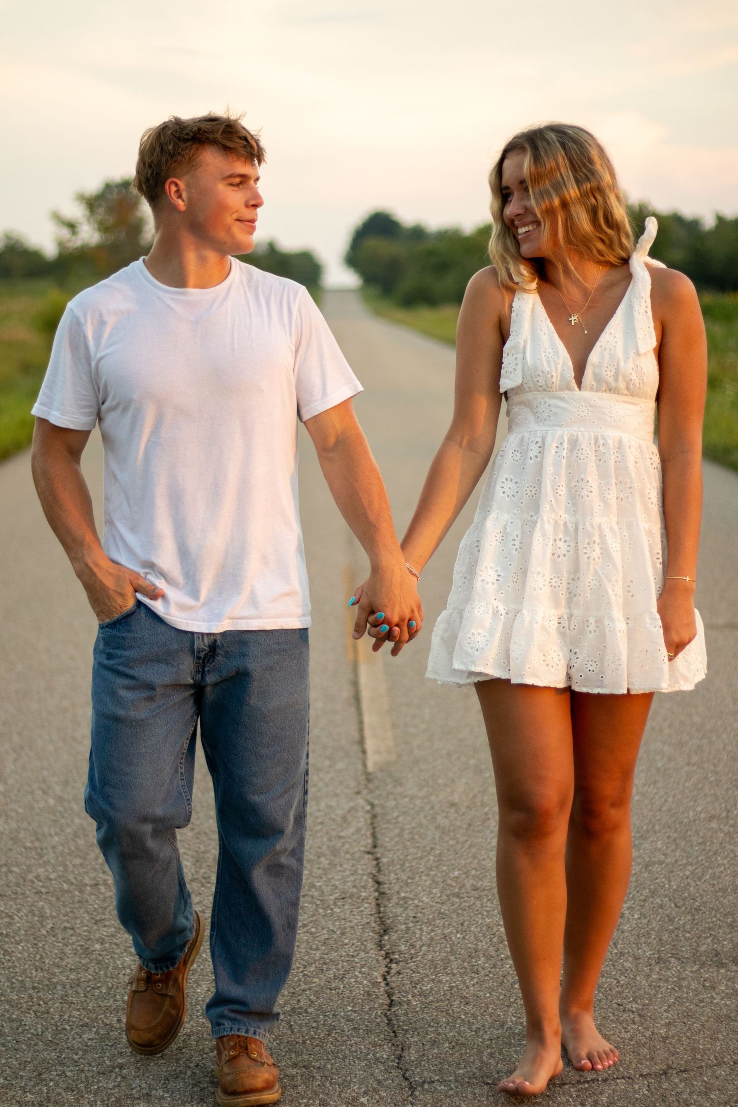 Happy couple holding hands and walking barefoot on an empty road at sunset smiling at each other.