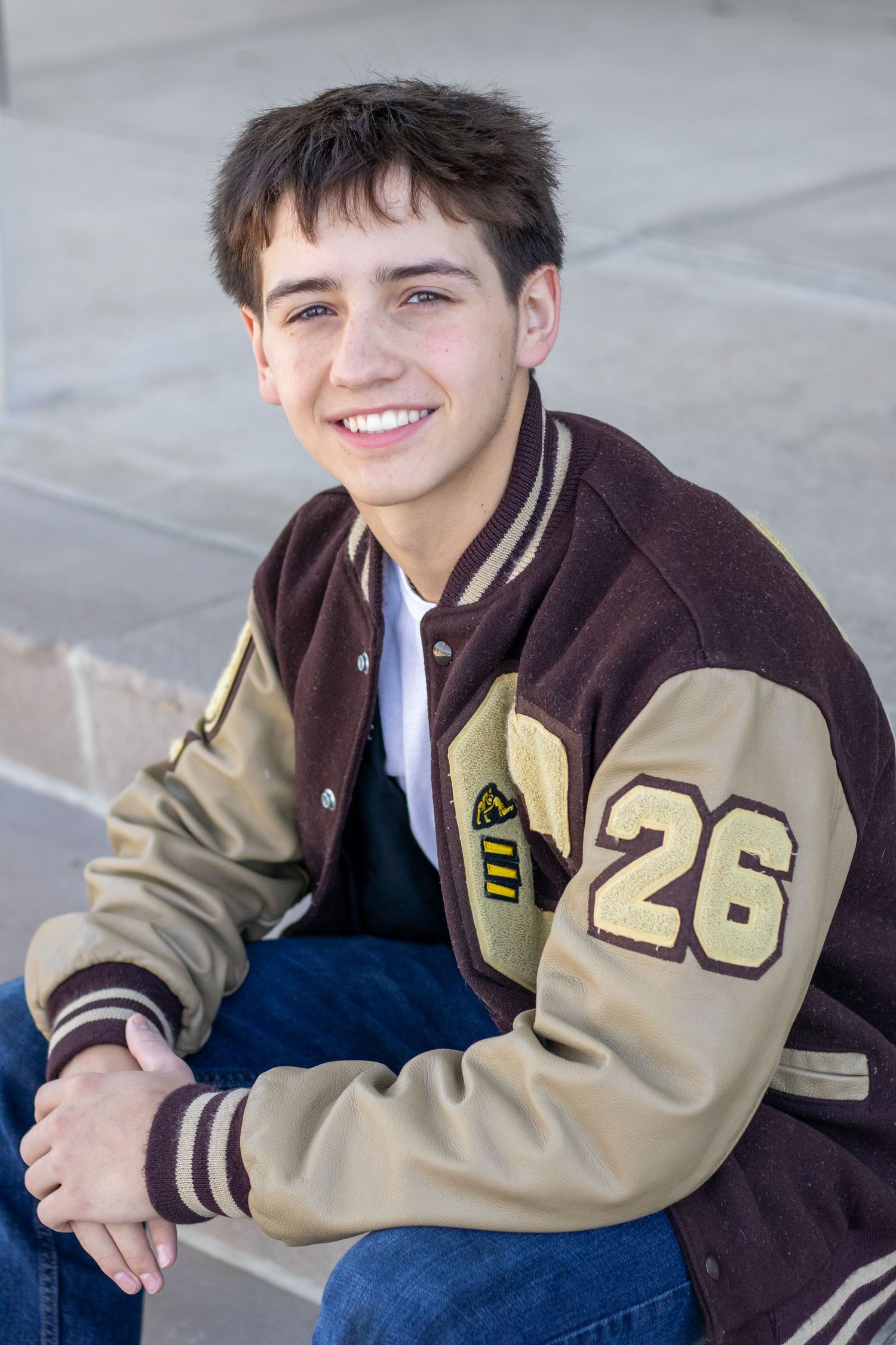 Happy teenage boy in brown and gold letterman jacket number 26 sitting outdoors smiling.