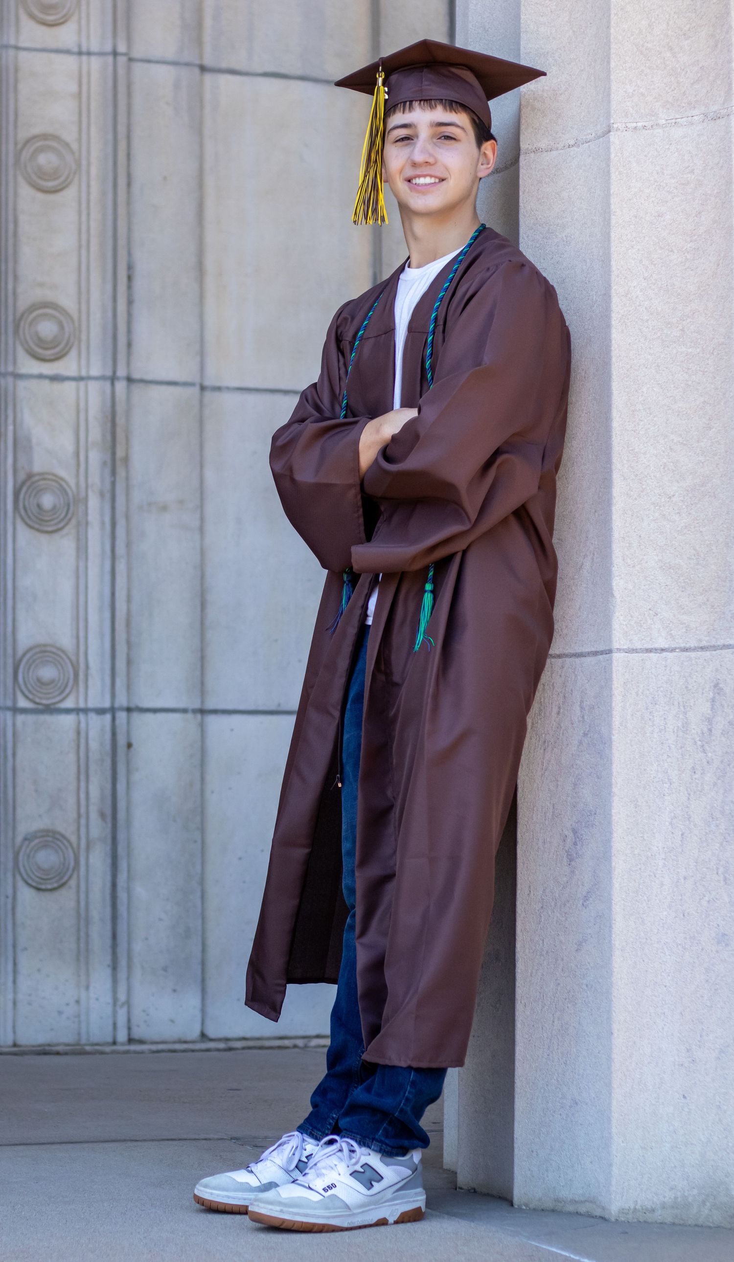 Male high school graduate in brown cap and gown leaning against stone column senior portrait.