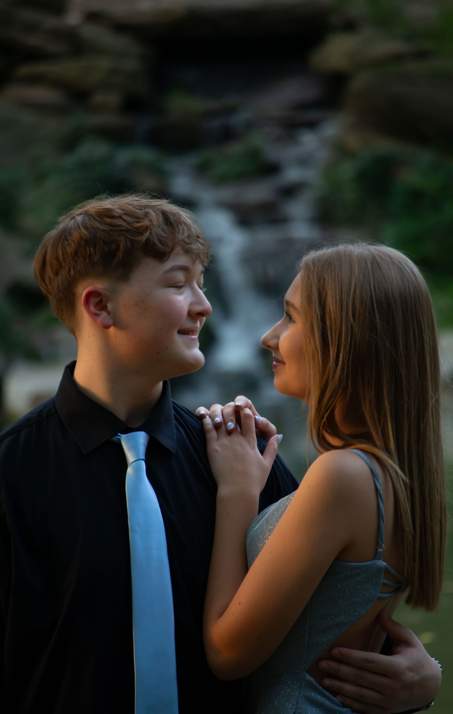 Teen couple smiling at each other in front of waterfall, boy in blue tie.