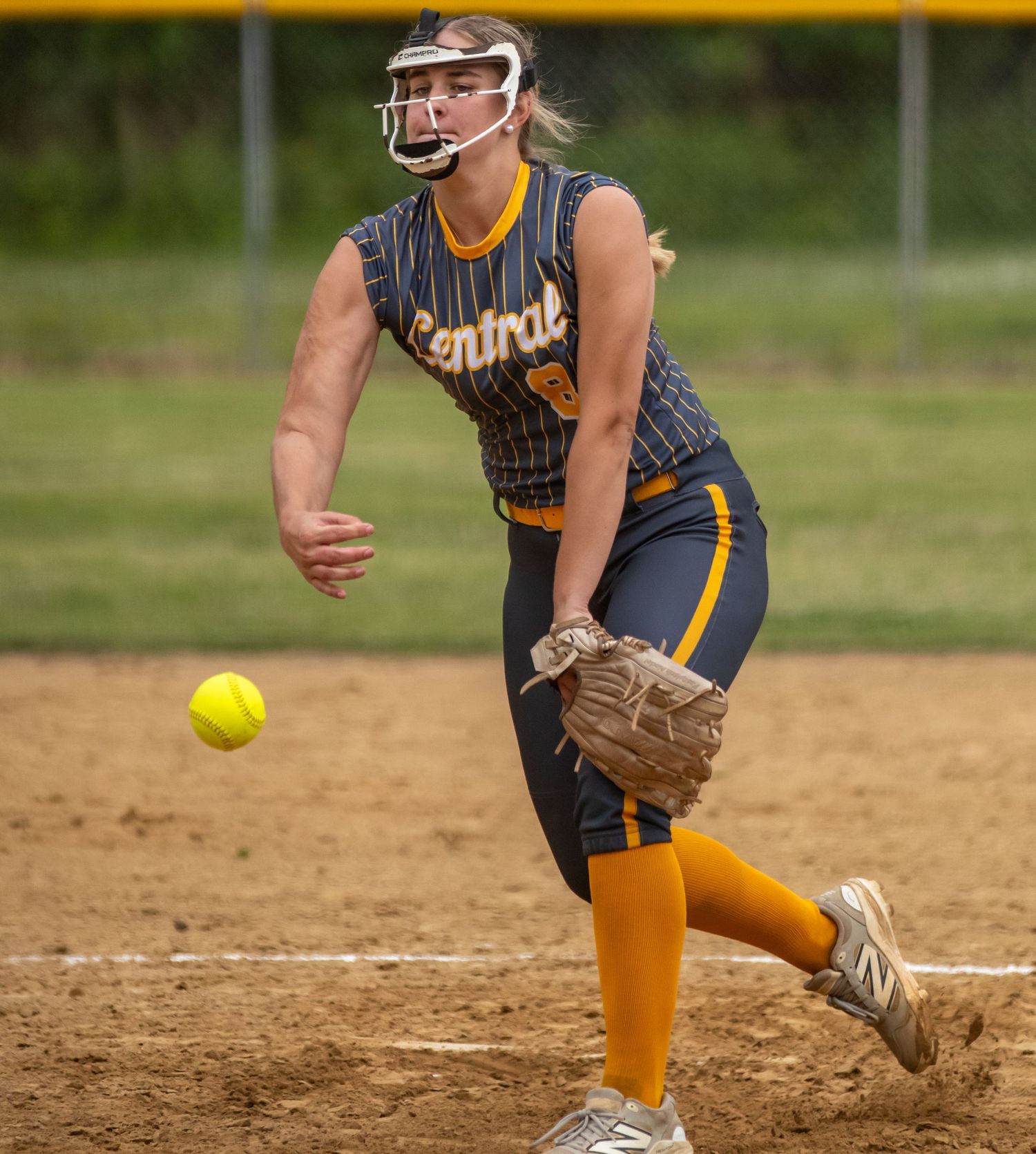 Female softball pitcher in Central uniform mid-throw, releasing yellow ball during competitive game outdoors.