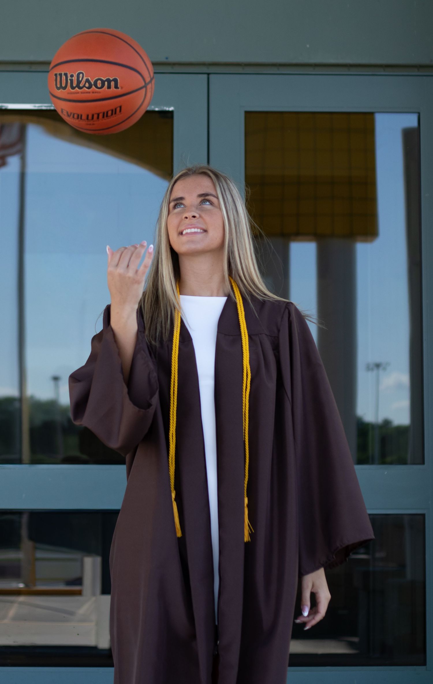 Blonde graduate in brown gown tossing a Wilson basketball outside school building.