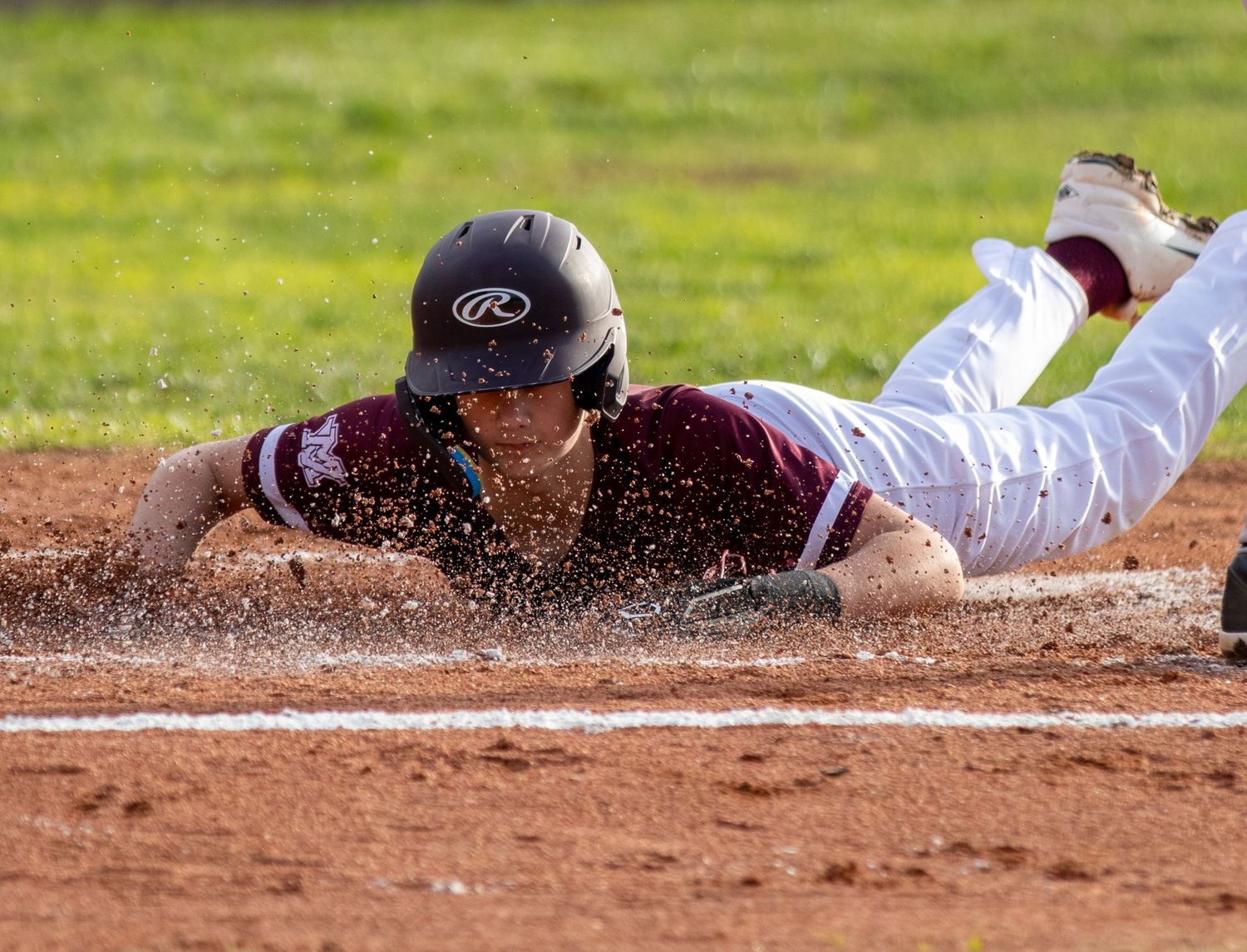 Baseball player in maroon uniform diving headfirst into base in a cloud of dirt.